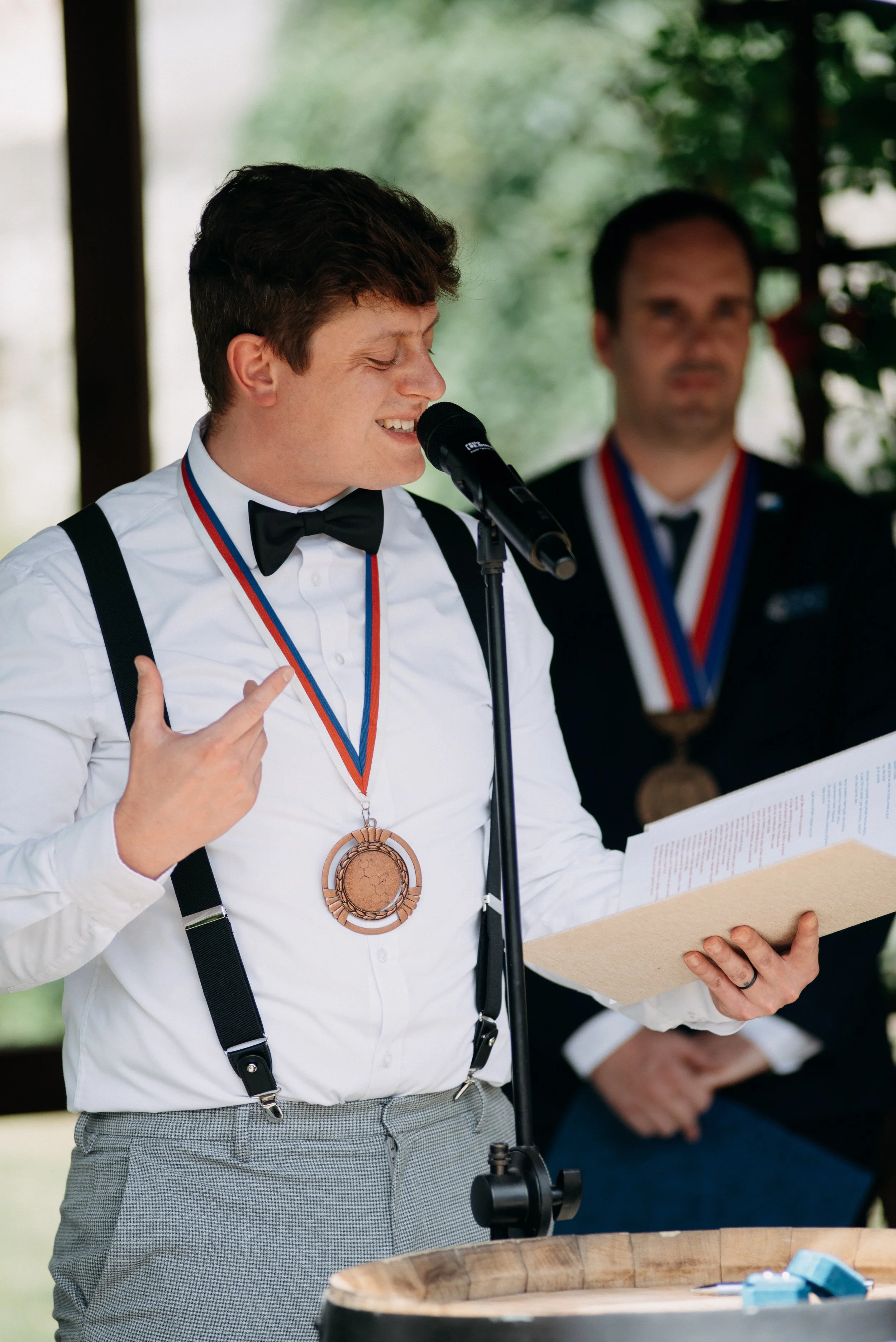 Oddávající Tukan v bílé košili, černých šlech a motýlku mluví do mikrofonu během svatebního obřadu. Za ním stojí starosta v obleku s medailí a v ruce drží dokument.