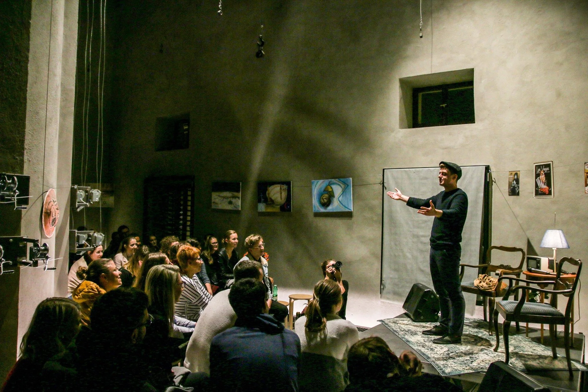 Slamer Tukan vystupuje na jevišti před publikem v místnosti s minimalistickou výzdobou, včetně židlí, lampy a uměleckých děl na stěnách. Slam poetry event.
