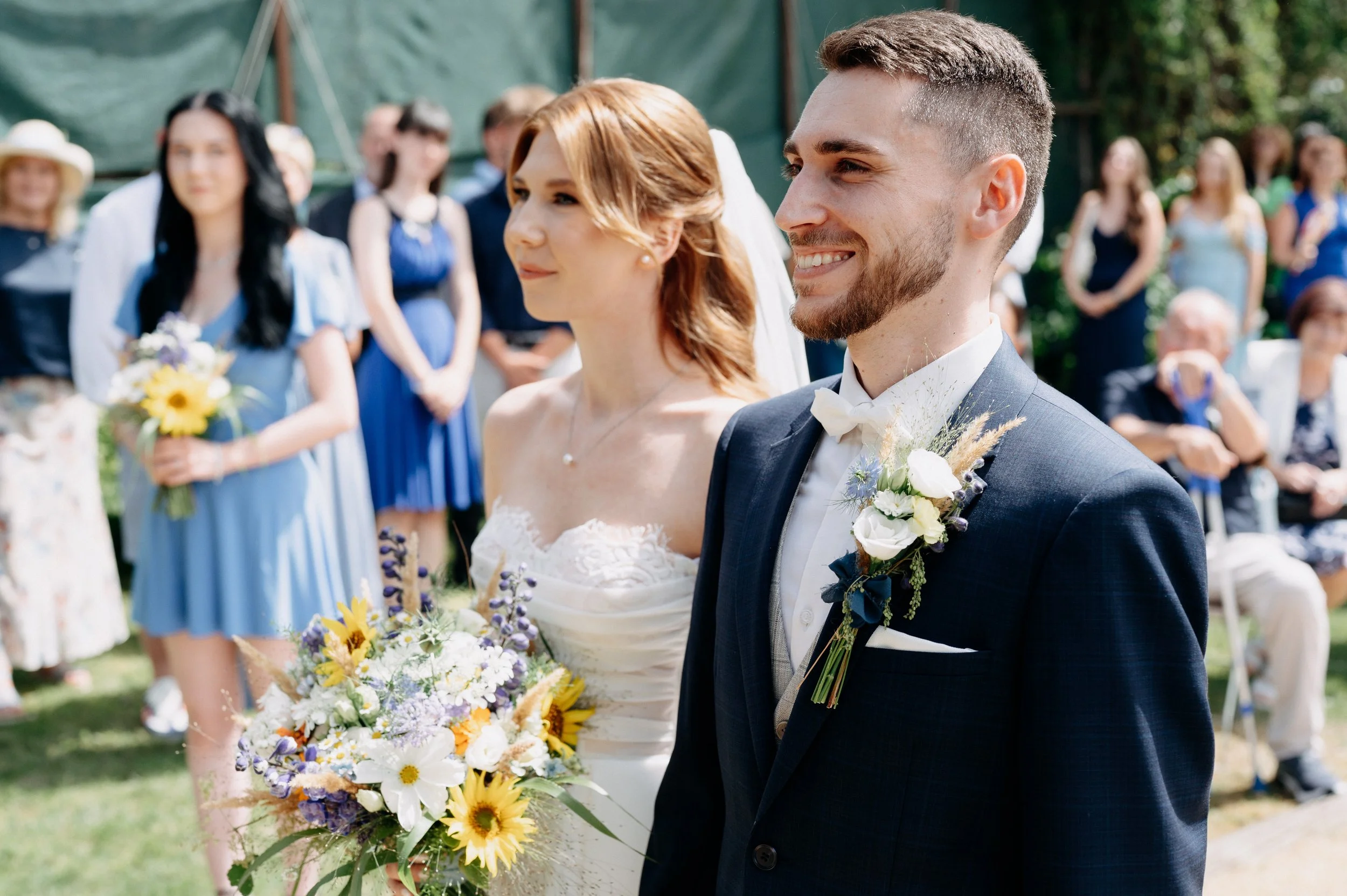 Nevěsta a ženich stojící venku během svatebního obřadu, s hosty v pozadí.