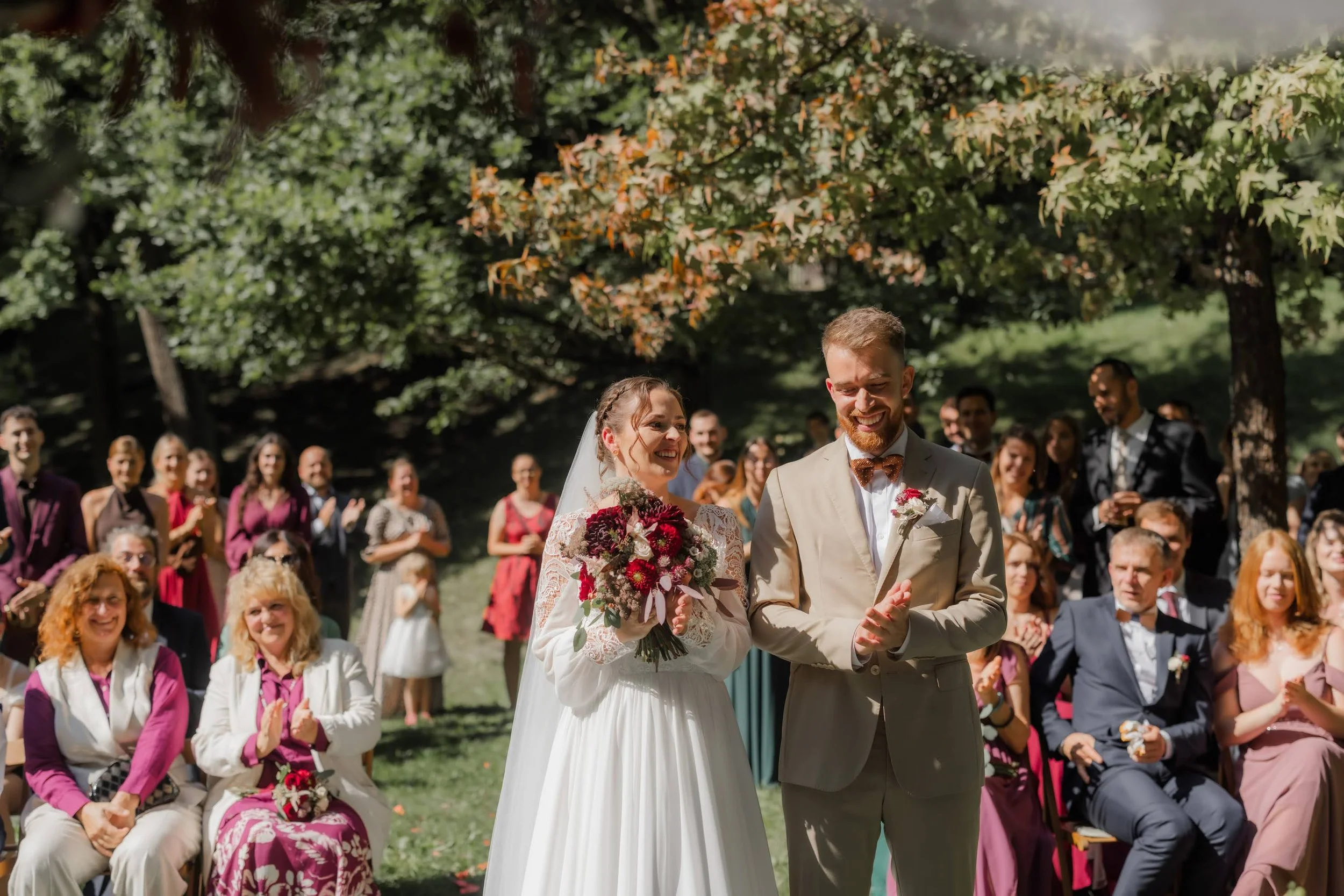 Nevěsta a ženich se usmívají na svém svatebním obřadu venku pod stromy, hosté sedí a stojí v pozadí a tleskají.