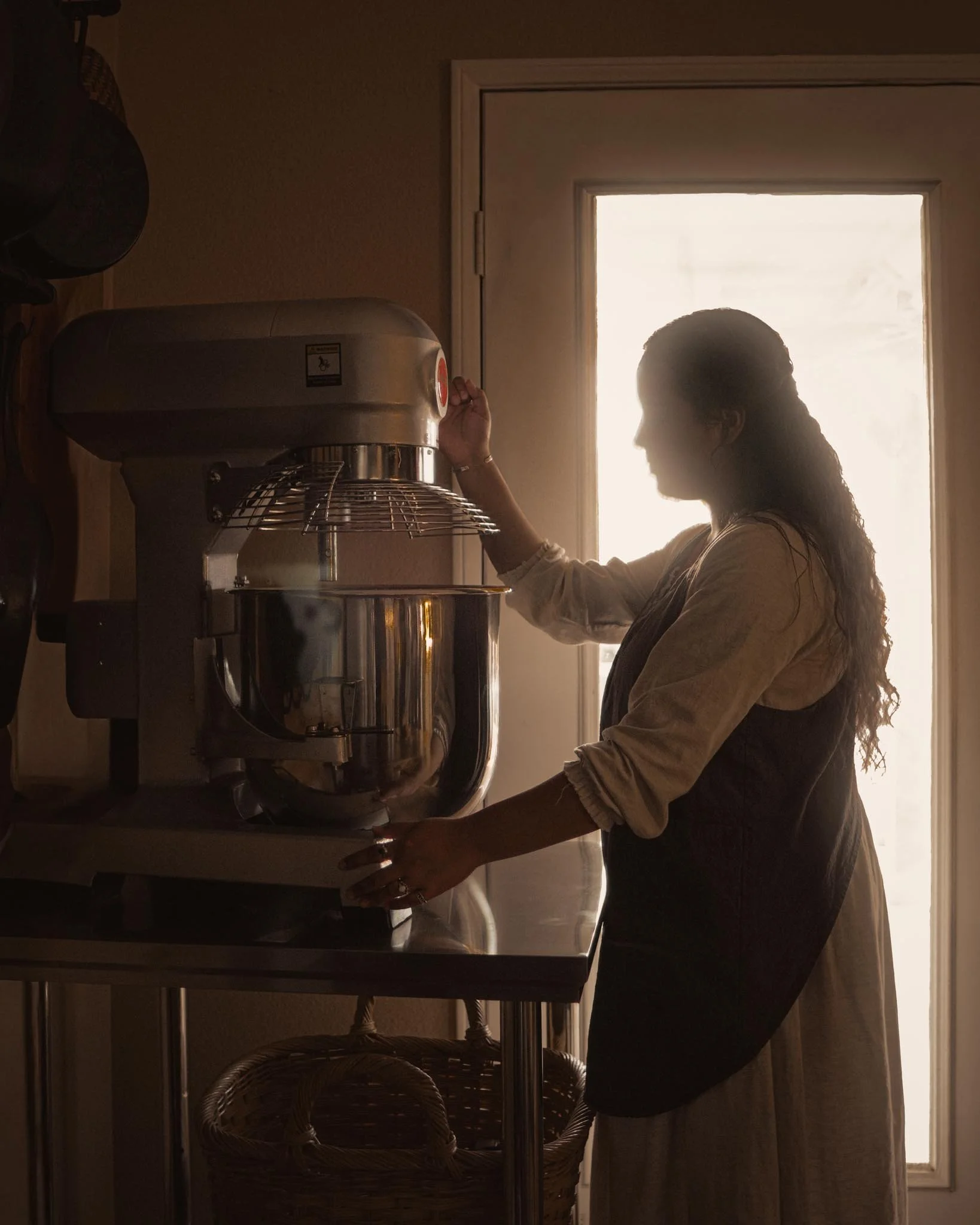 A woman standing near a stand mixer in a kitchen, adjusting it, with a window in the background letting in natural light.