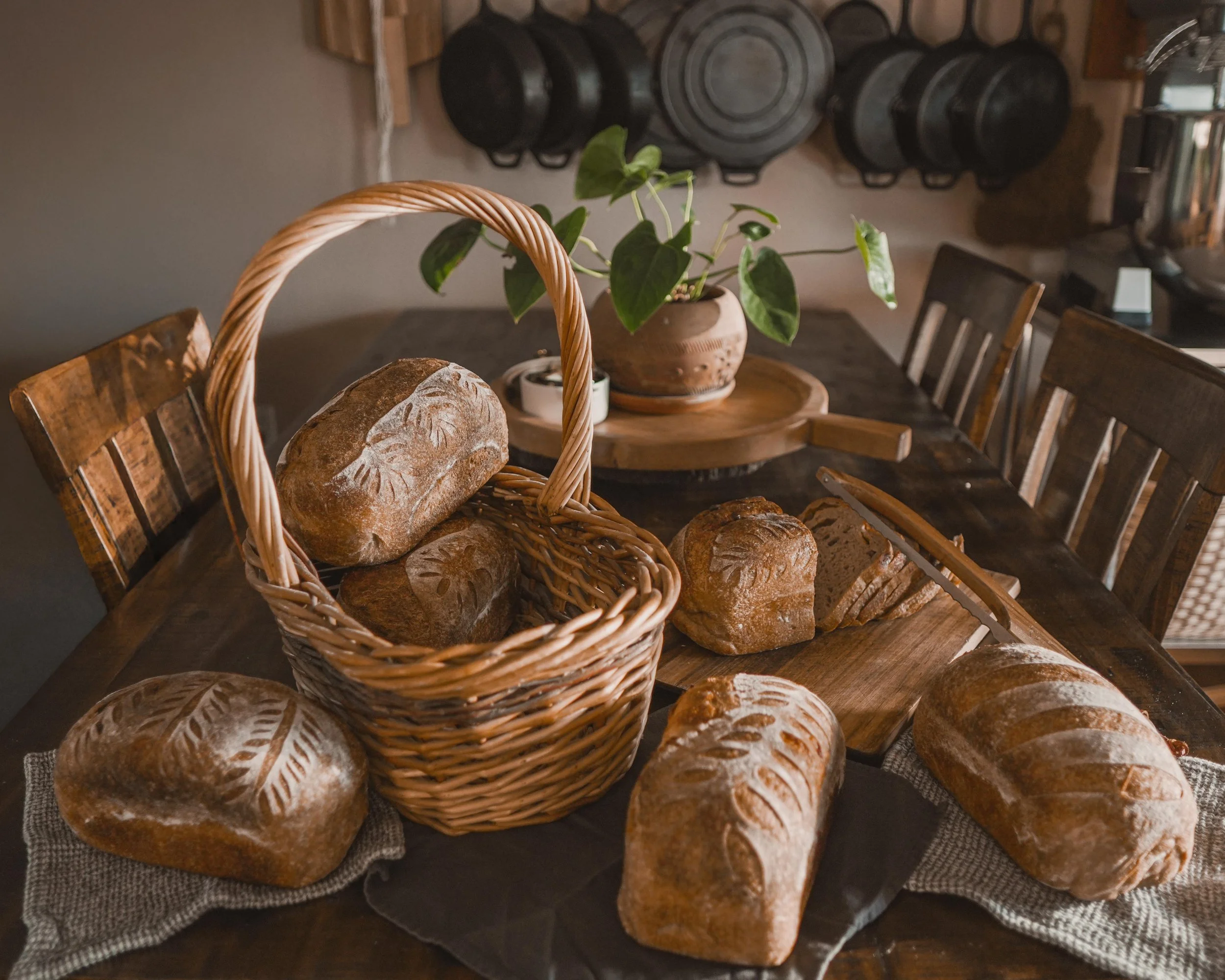 A wooden dining table with various loaves of bread, some on a cloth and others in a wicker basket. A potted plant and a round wooden tray are in the background, with hanging pots on the wall.