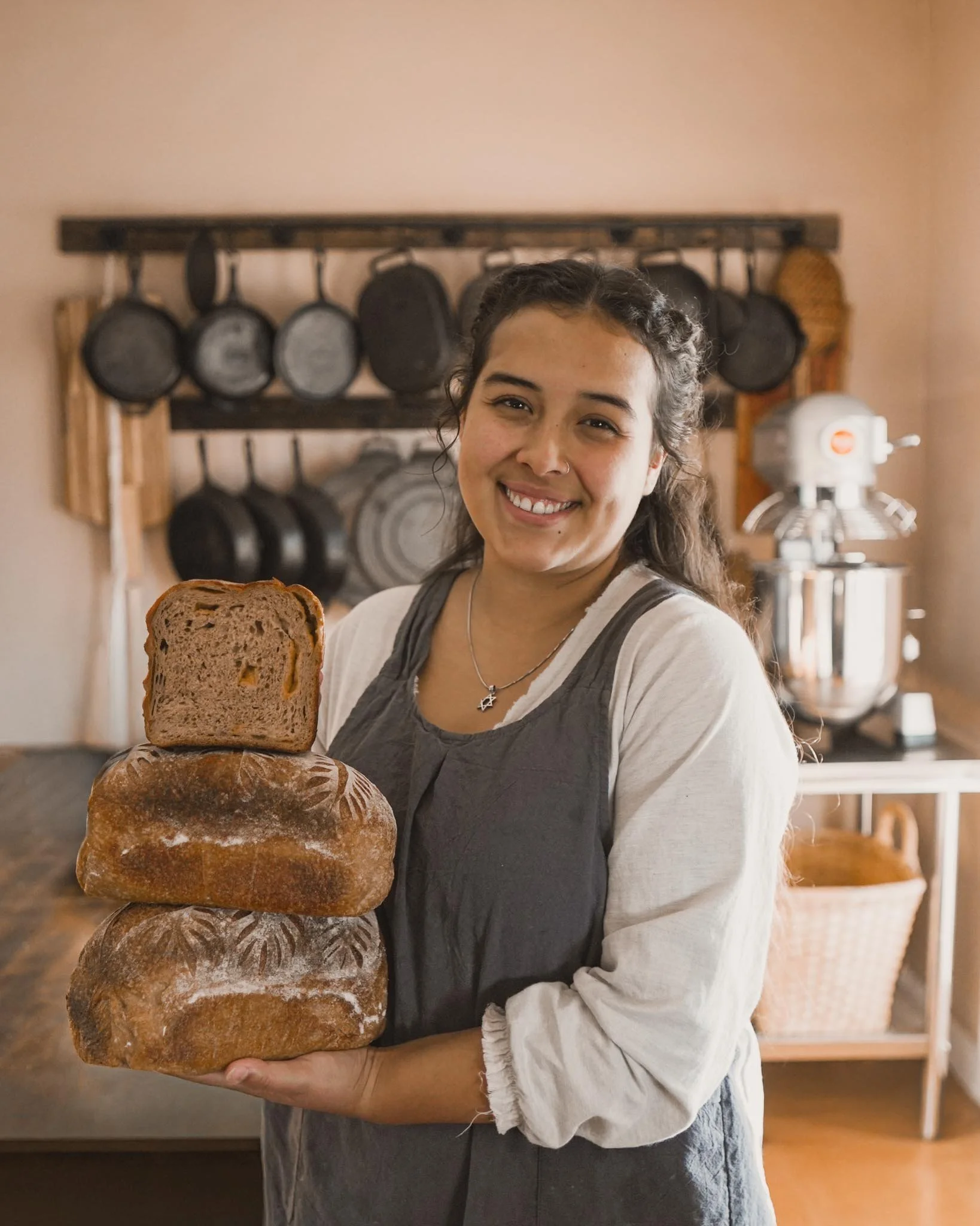 A woman smiling and holding a stack of three loaves of bread in a kitchen with hanging cast iron skillets in the background.