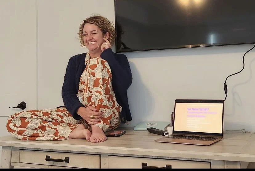 Woman sitting cross-legged on a table, smiling, with a TV behind her and a laptop on the table displaying a presentation.