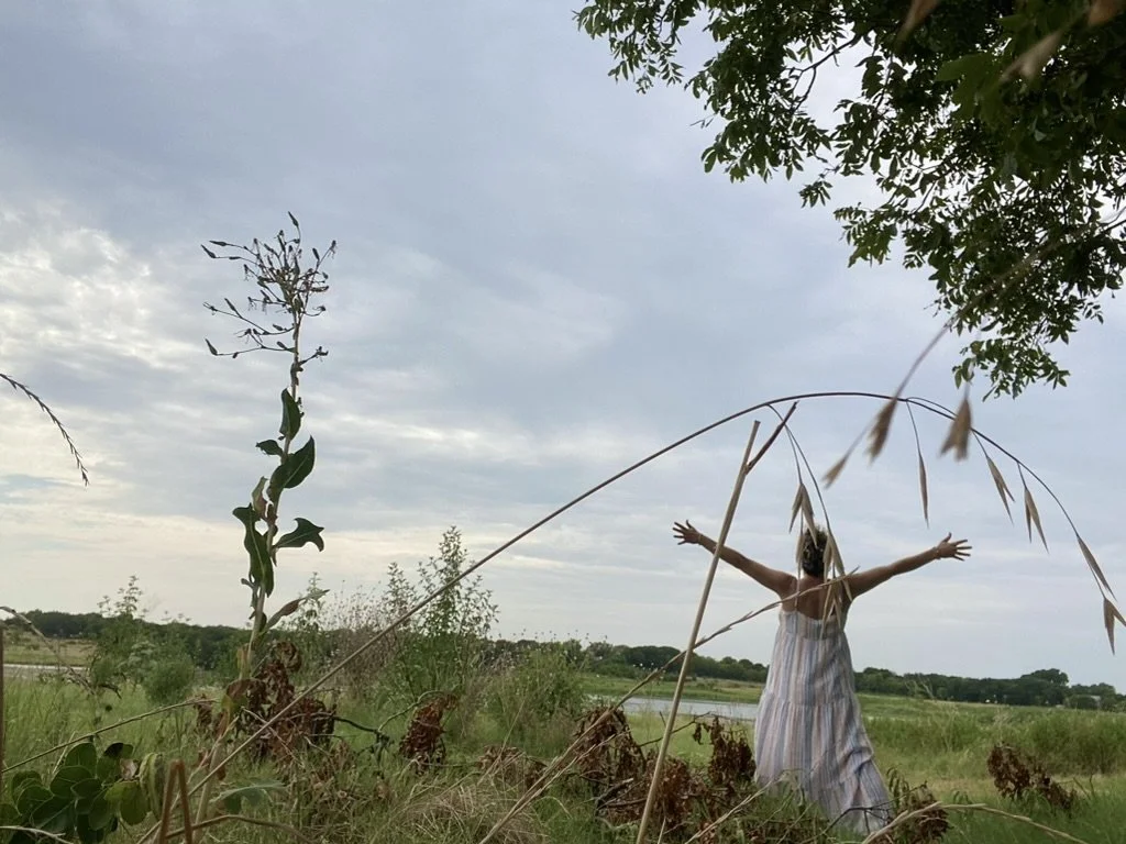 A woman standing outside in a field with her arms outstretched, wearing a striped dress and surrounded by tall grass, plants, and trees under a cloudy sky.