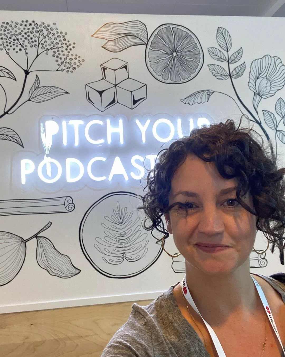 A woman taking a selfie in front of a wall with the neon sign 'PITCH YOUR PODCAST' and blackline illustrations of leaves, a citrus fruit slice, cubes, and a fern.