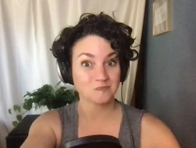A woman wearing headphones taking a selfie in a room with a plant and a curtain in the background.