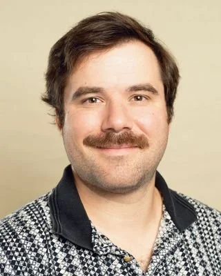 Man with brown hair and mustache wearing a polo shirt with a black and white print smiles at the camera