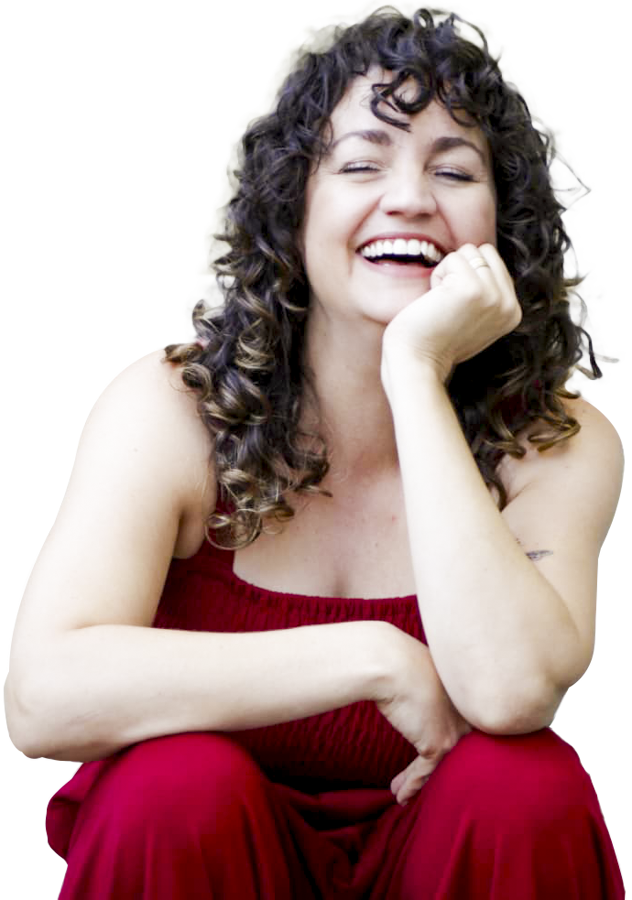 A woman with curly brown hair, smiling and laughing, sitting with her hand resting on her chin, wearing a red sleeveless top and red pants.