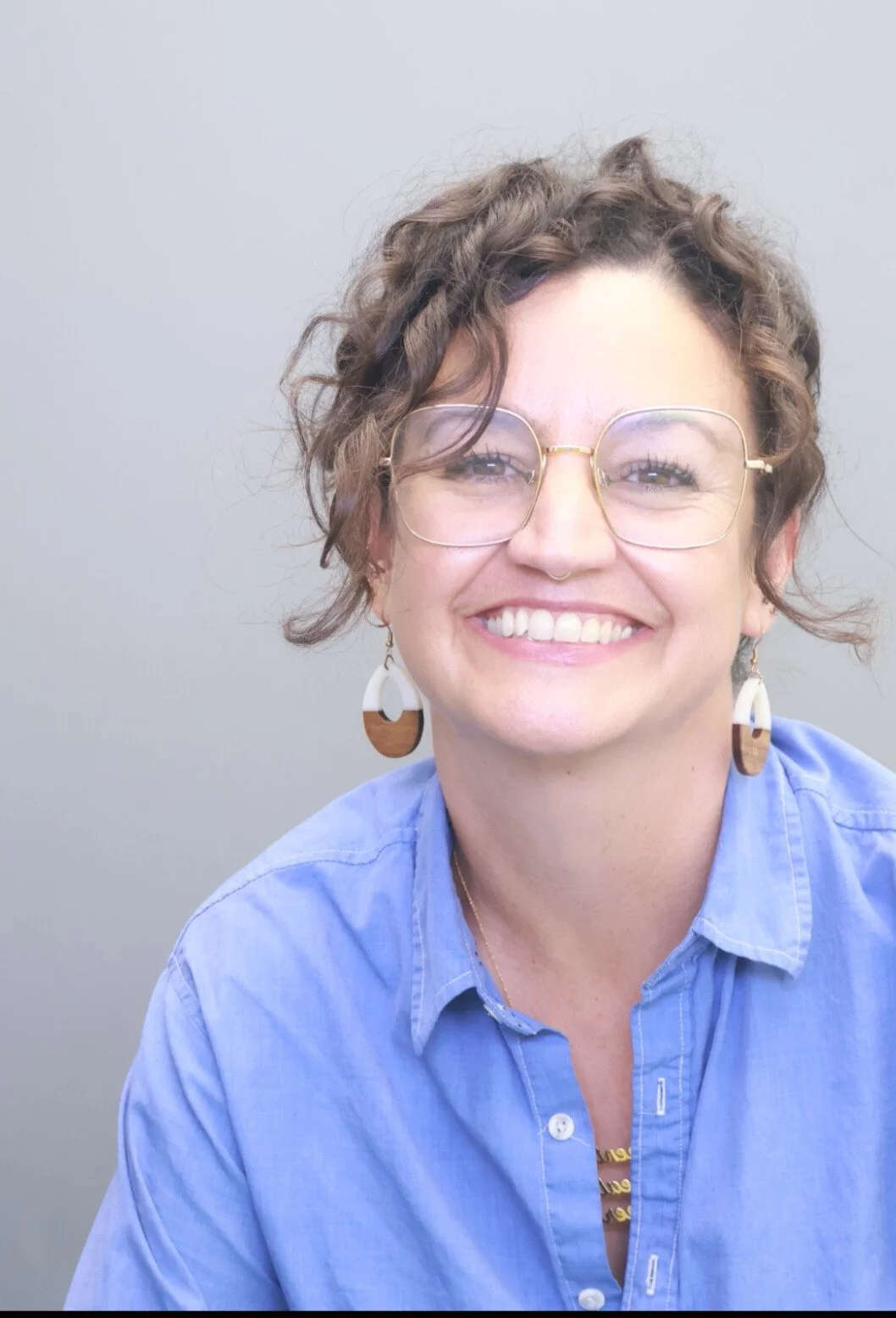 Woman with brown curly hair wearing earrings, necklace, and a blue button down shirt smiles at the camera