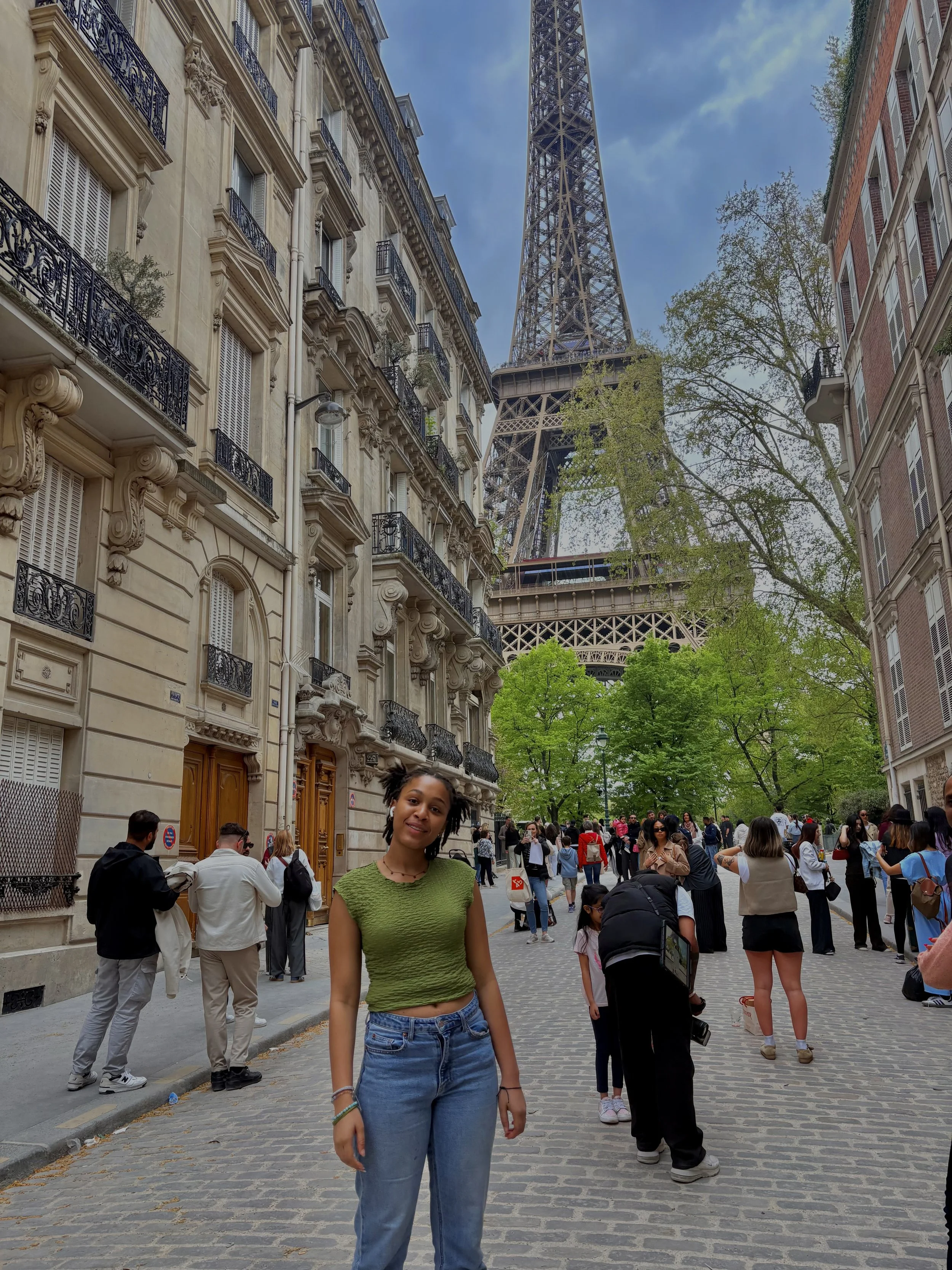 A young woman with short black hair, wearing a green textured crop top and blue jeans, standing on a cobblestone street in front of the Eiffel Tower in Paris, France, with several people around and trees lining the street.