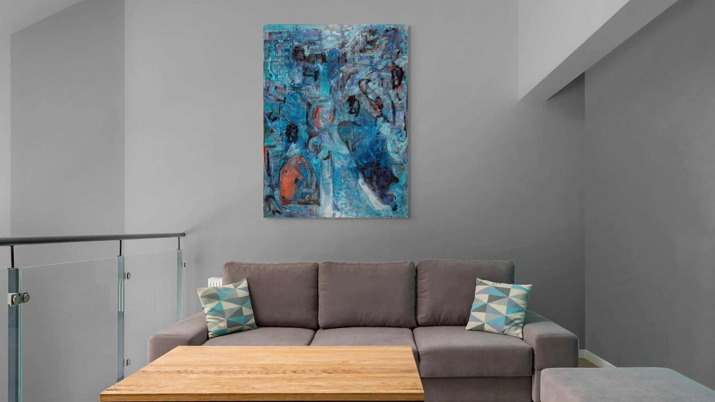 modern living room with a minimalist design. A grey sectional sofa with two patterned accent pillows sits against a light grey wall, above which hangs a vibrant abstract painting dominated by blue and teal tones with some orange accents. Overall, the