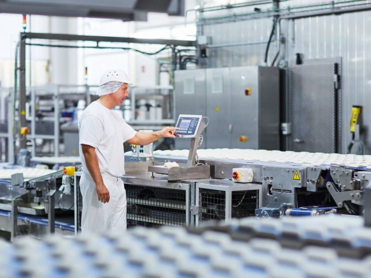 A worker in a white uniform and hairnet operating a control panel in a food manufacturing facility with production lines and machinery in the background.