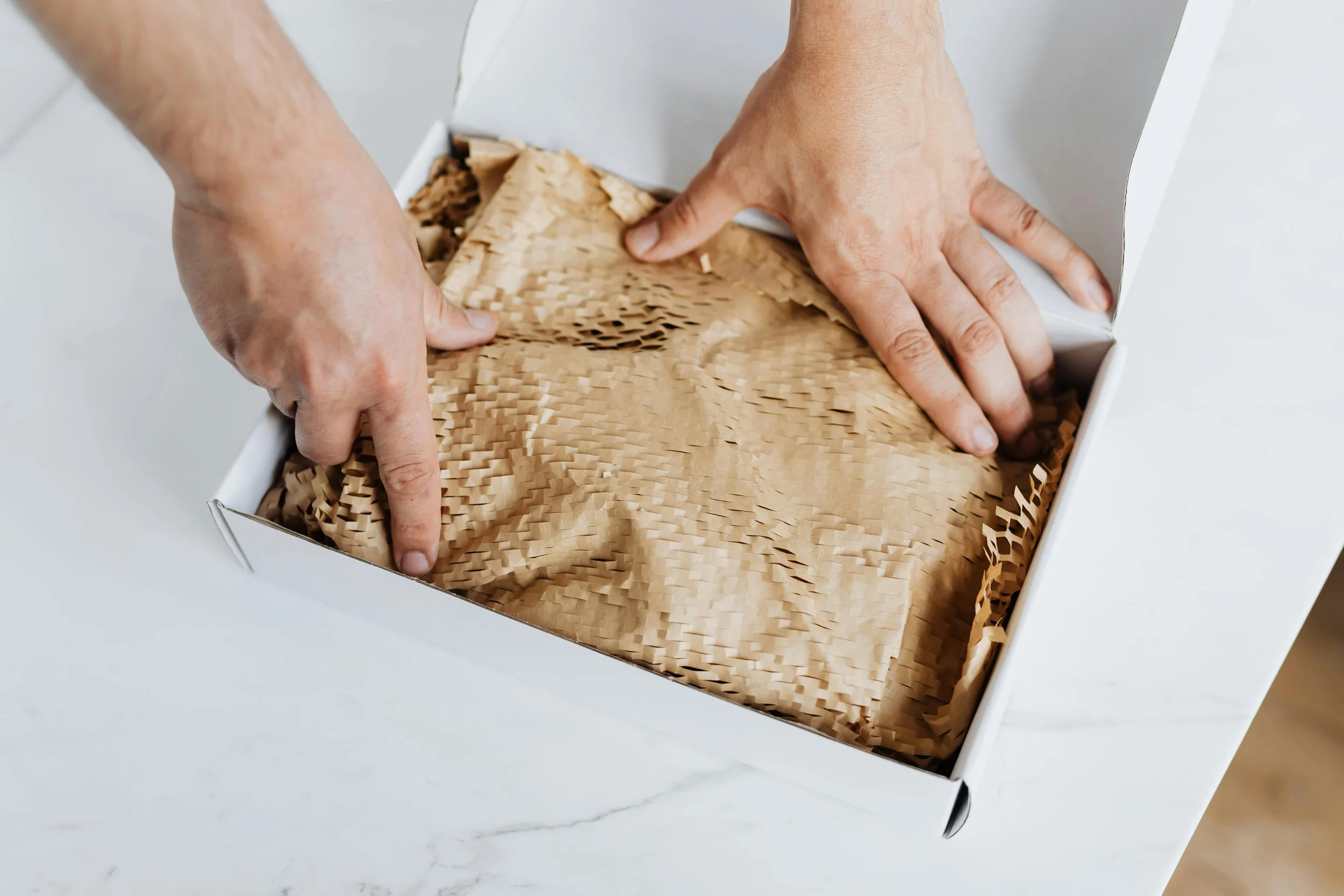 Hands opening a cardboard box filled with crumpled paper packaging material.