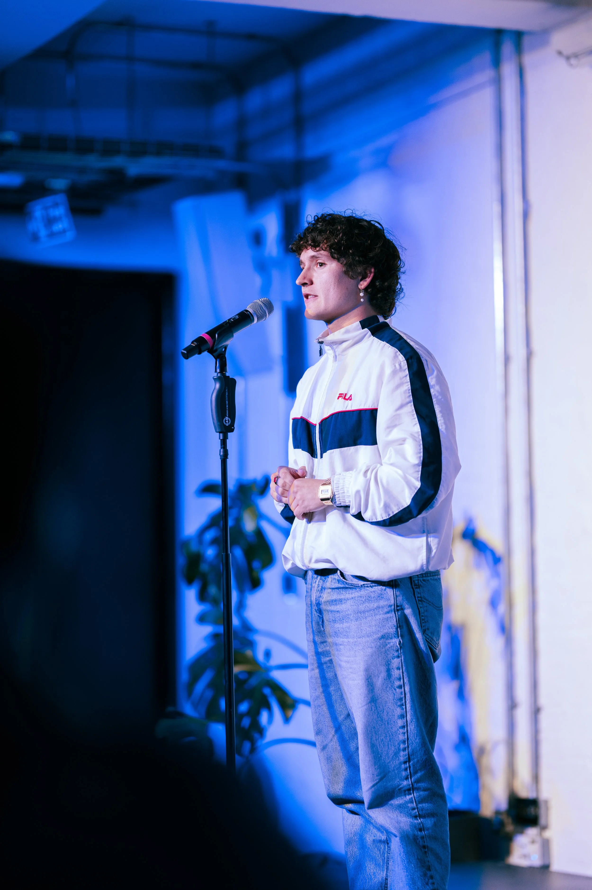 A young man with curly hair wearing a white Fila jacket and jeans stands on stage, speaking into a microphone, with blue lighting and a plant in the background.