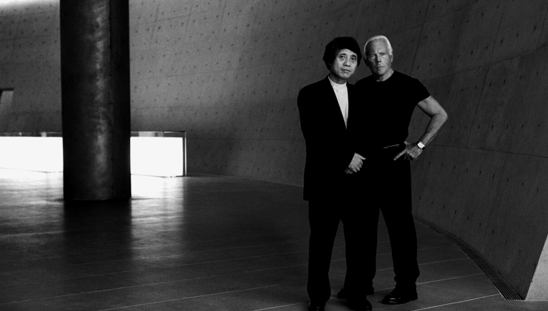 Two men standing side by side in front of a curved wall in a modern, minimalist interior.