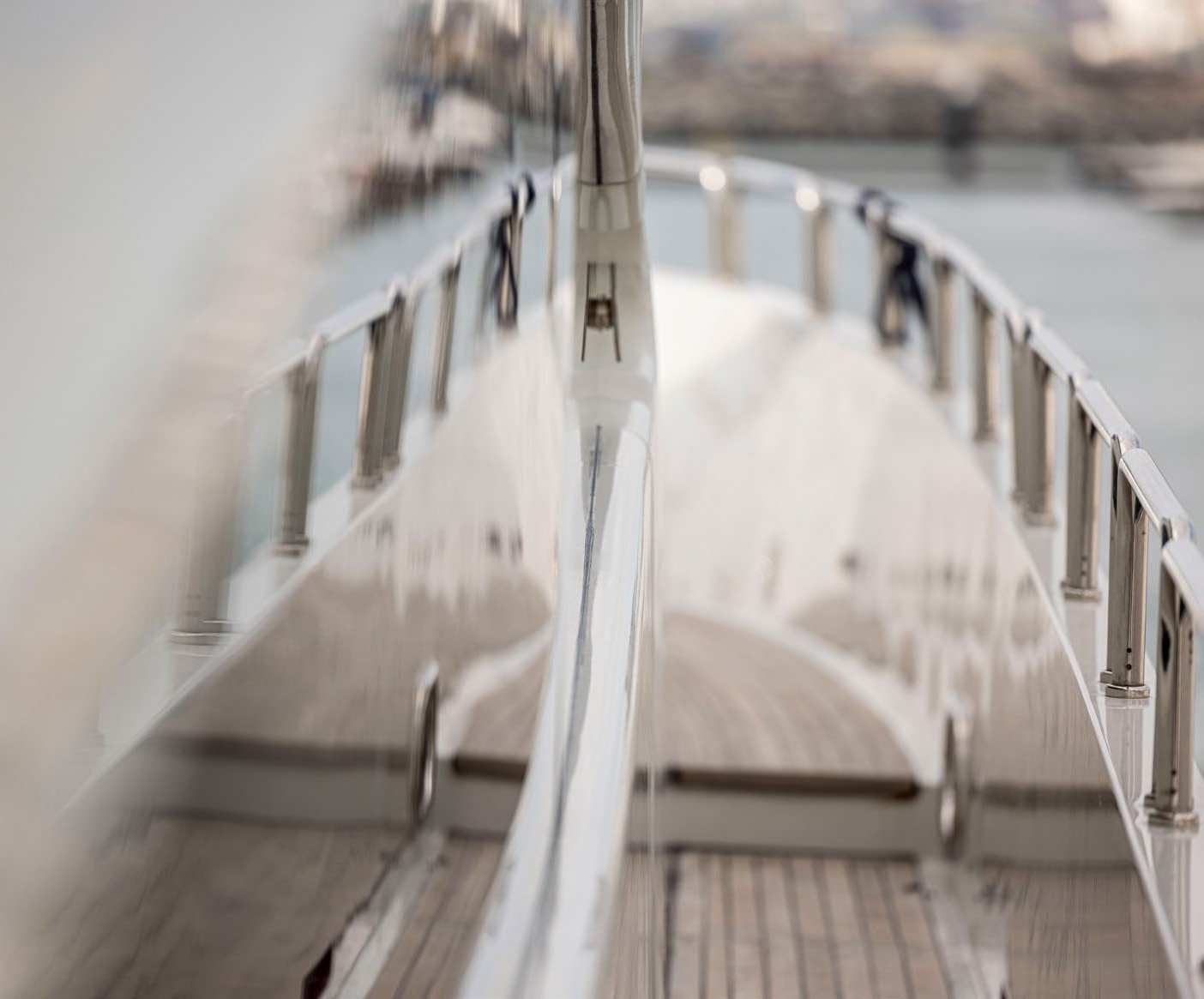 A close-up view of the bow of a yacht, showing stainless steel railings and a sleek white hull, with water and blurred cityscape in the background.