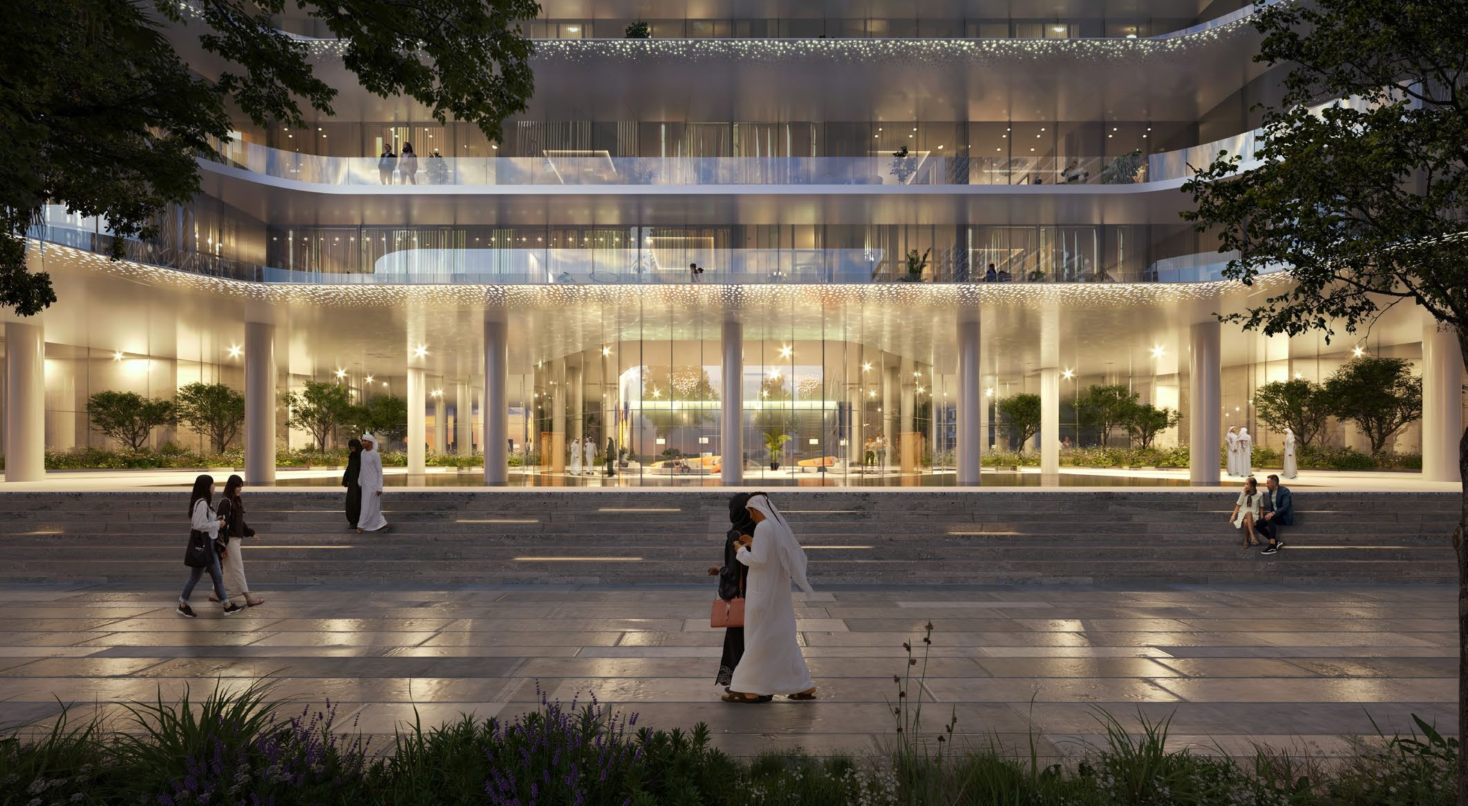 Exterior view of a modern multi-story building at dusk with glass walls and illuminated interior, surrounded by trees and people walking, sitting, and chatting outside.