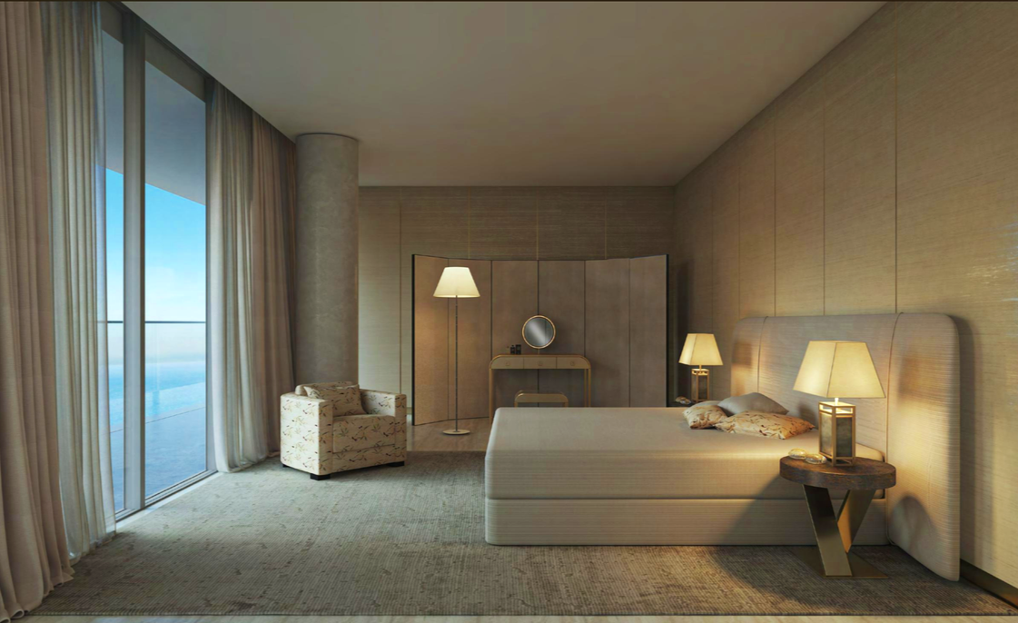 Hotel room with a large bed, bedside lamps, a floral armchair, a floor lamp, and a sliding glass door with beige curtains leading to a balcony with ocean view.