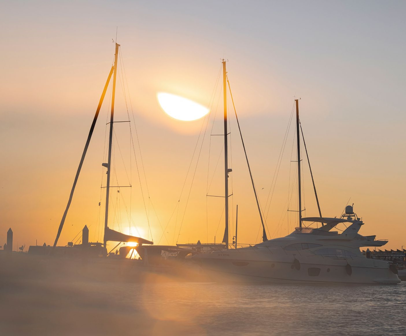 Sailboats and a yacht docked at a marina during sunset with the sun partially obscured by clouds