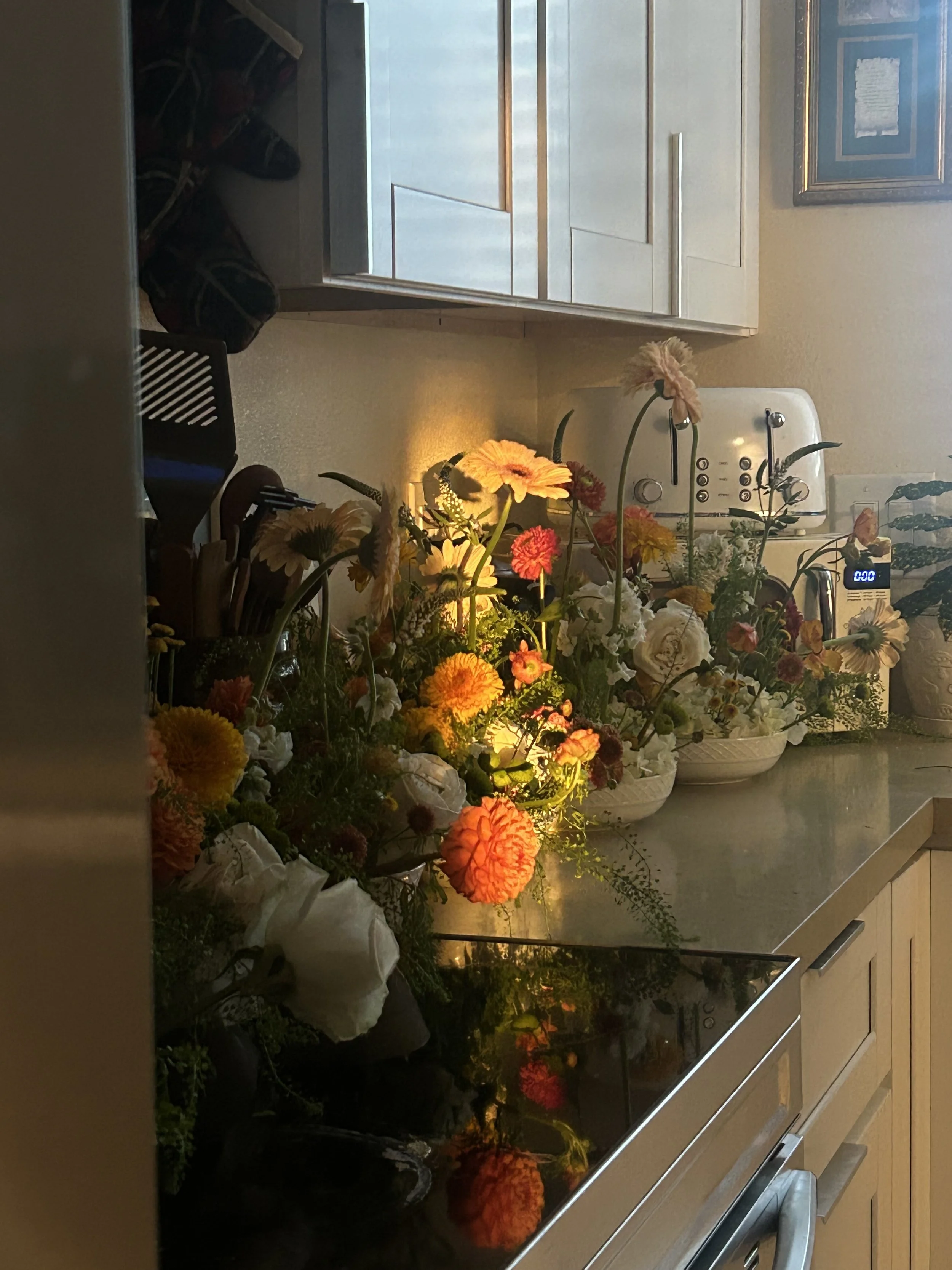 A kitchen countertop decorated with a variety of colorful flowers in bowls and vases, illuminated by a warm light, with cabinets, a toaster, and a digital clock in the background.
