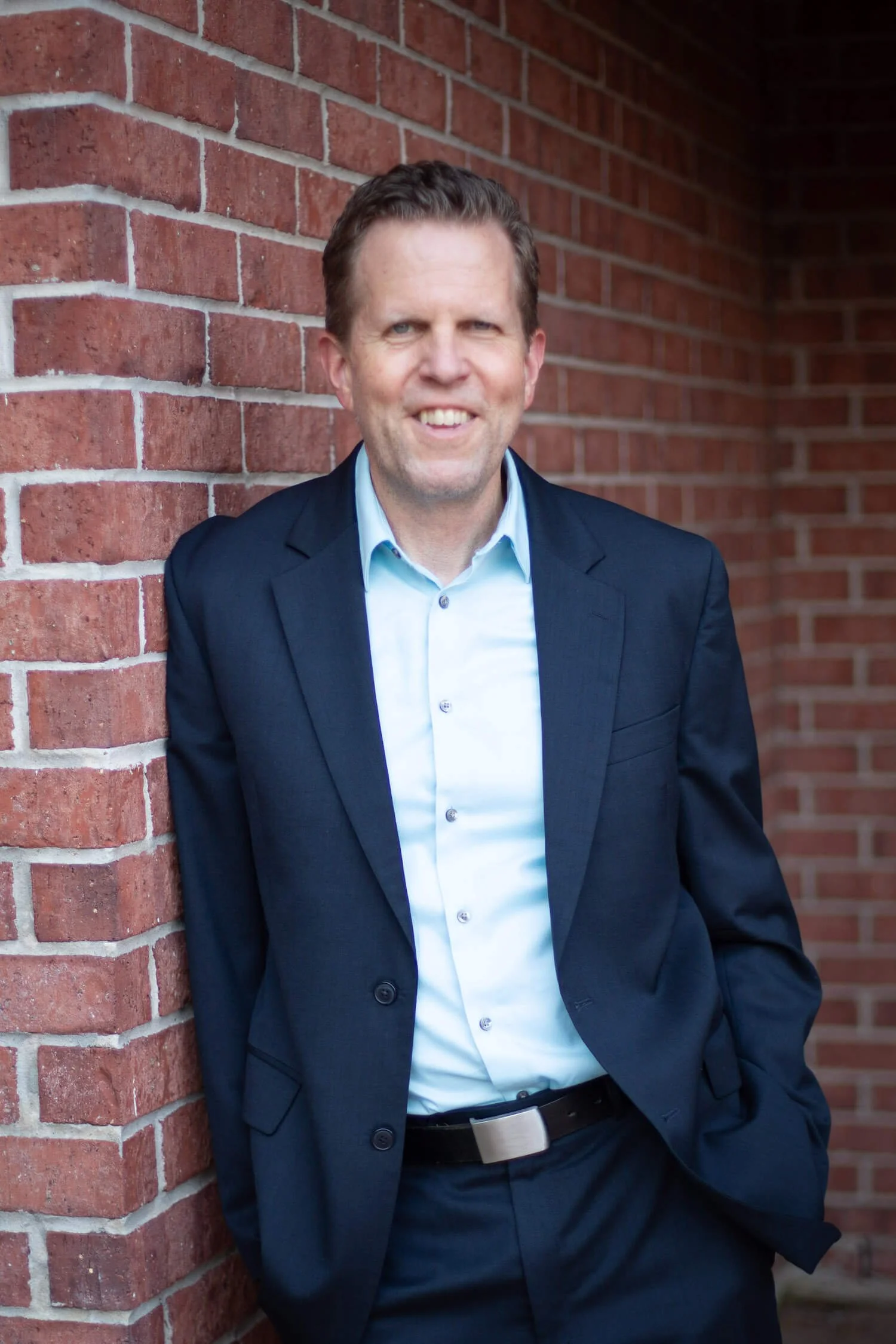 Executive coach Brian Owen in a blue shirt and dark blazer with arms crossed, leaning against a brick wall