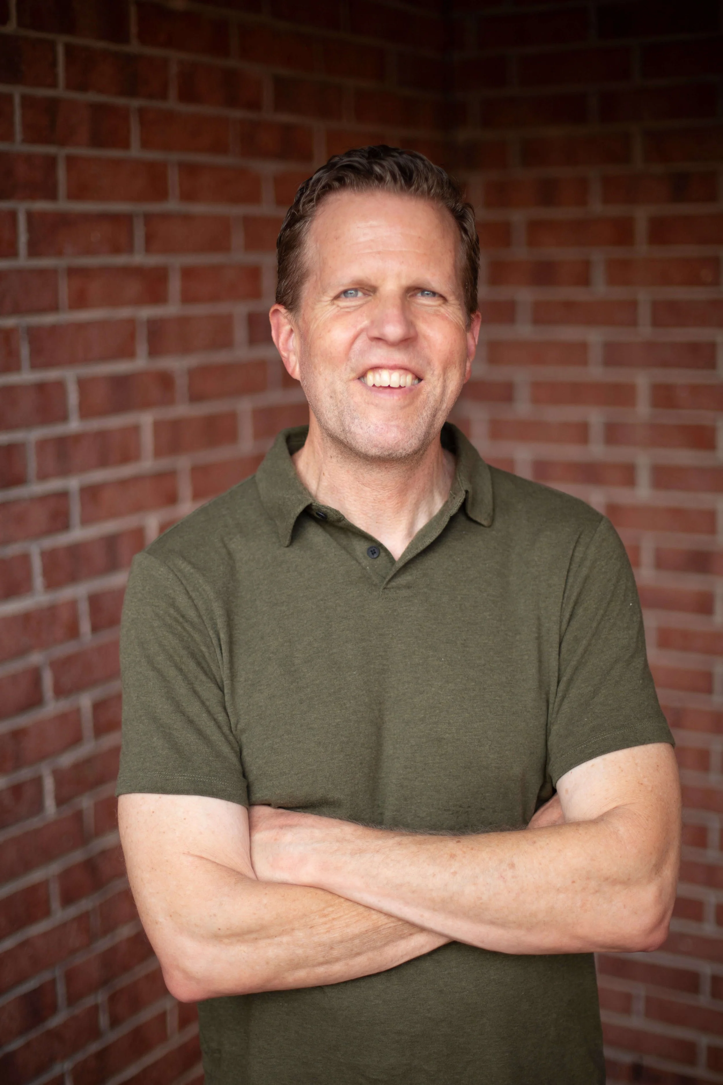 Executive coach Brian Owen, wearing a green polo shirt, standing with arms crossed in front of a red brick wall.