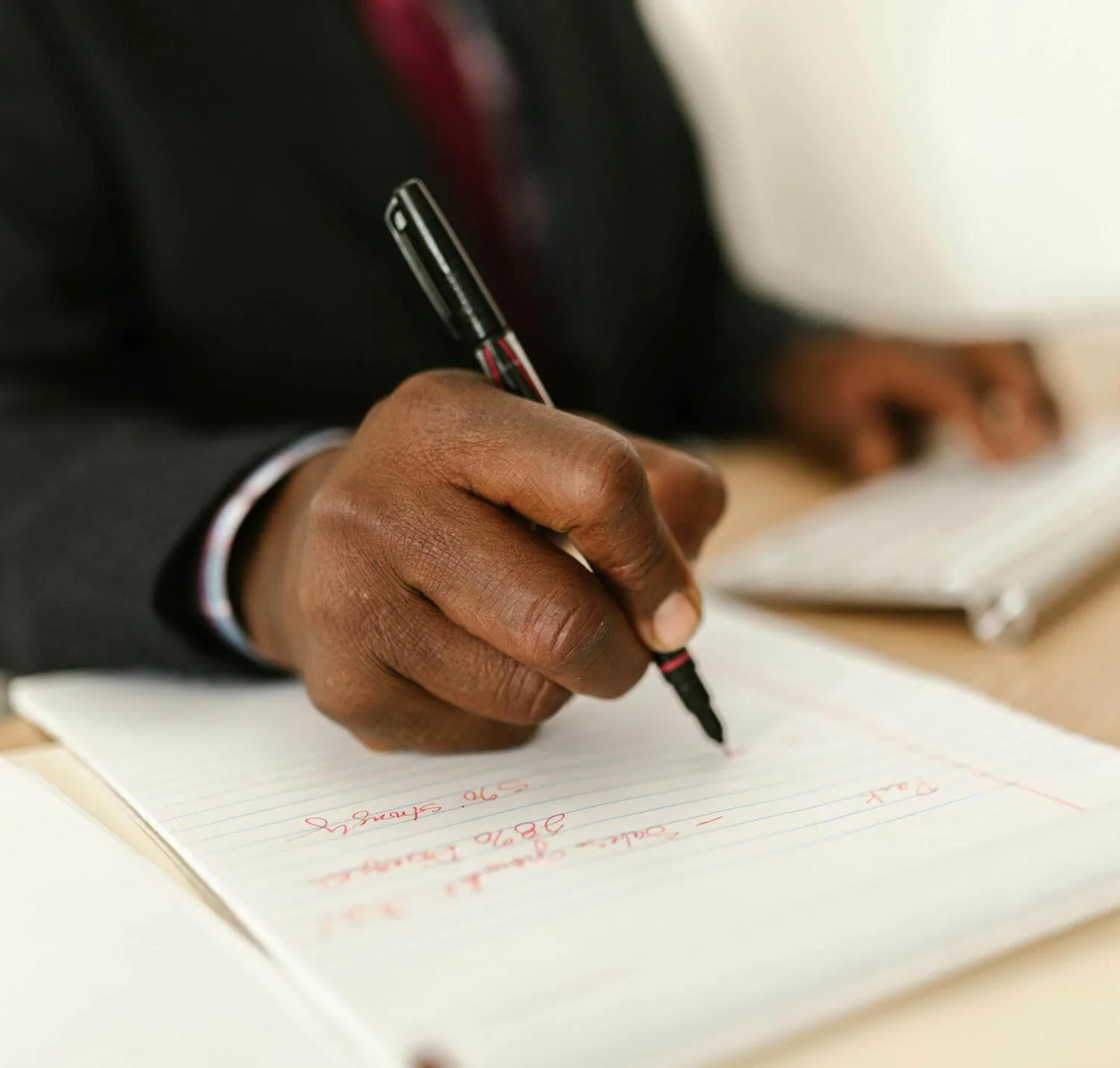 A person in a dark suit is writing in a notebook with a black pen with red accents.