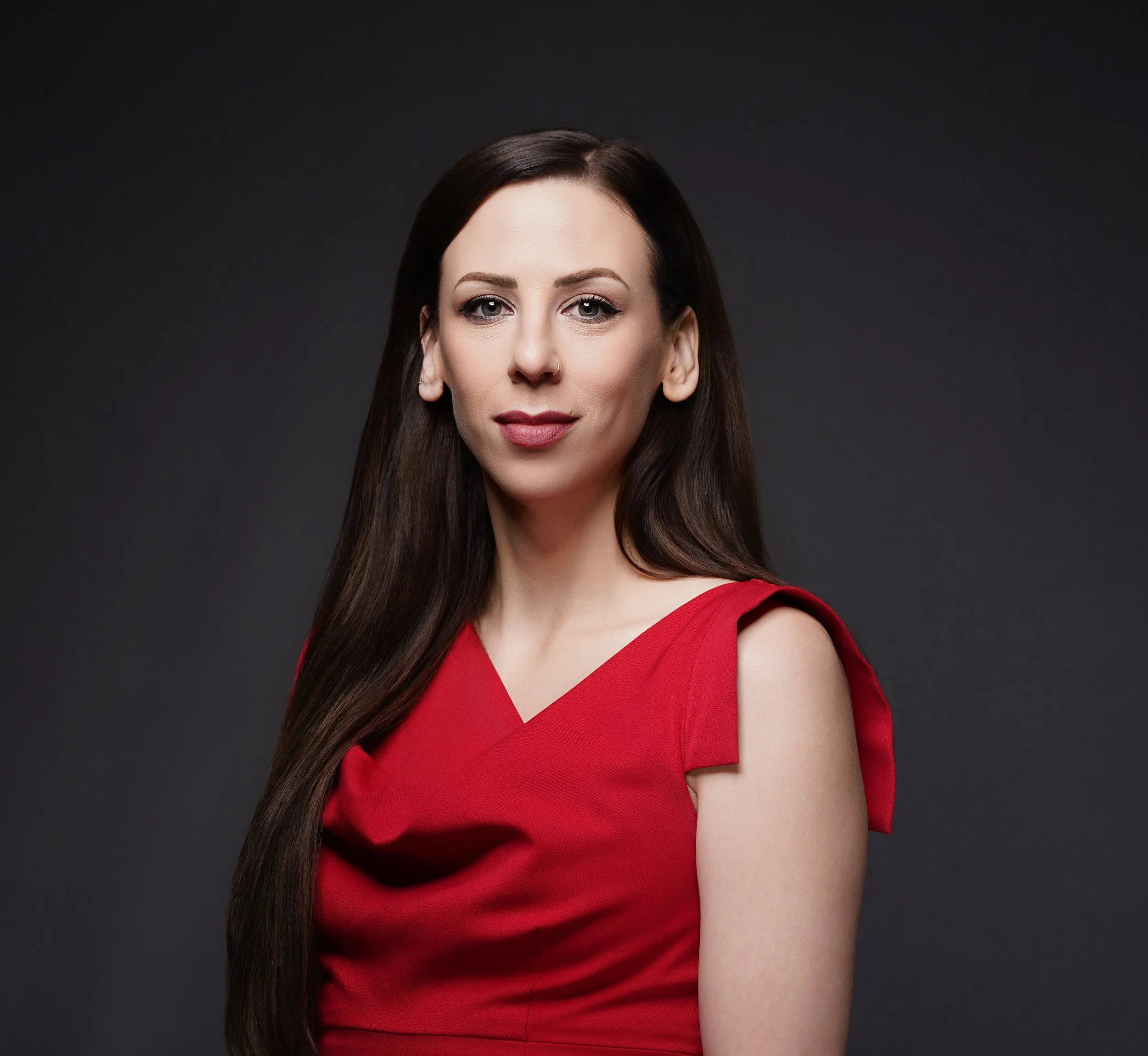 A woman with long brown hair wearing a red dress, standing against a dark background.