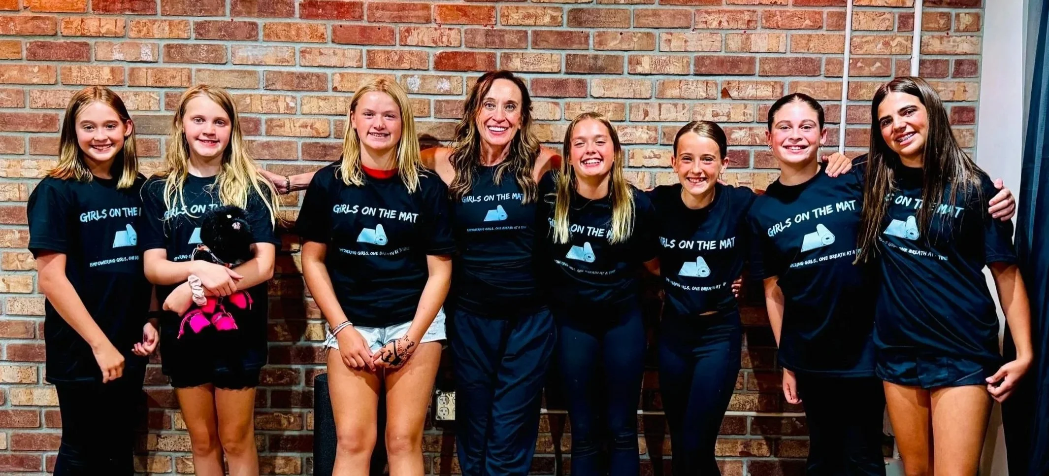 Group of eight girls standing together against a brick wall, all wearing matching black shirts that say "Girls on the Mat" and smiling at the camera.