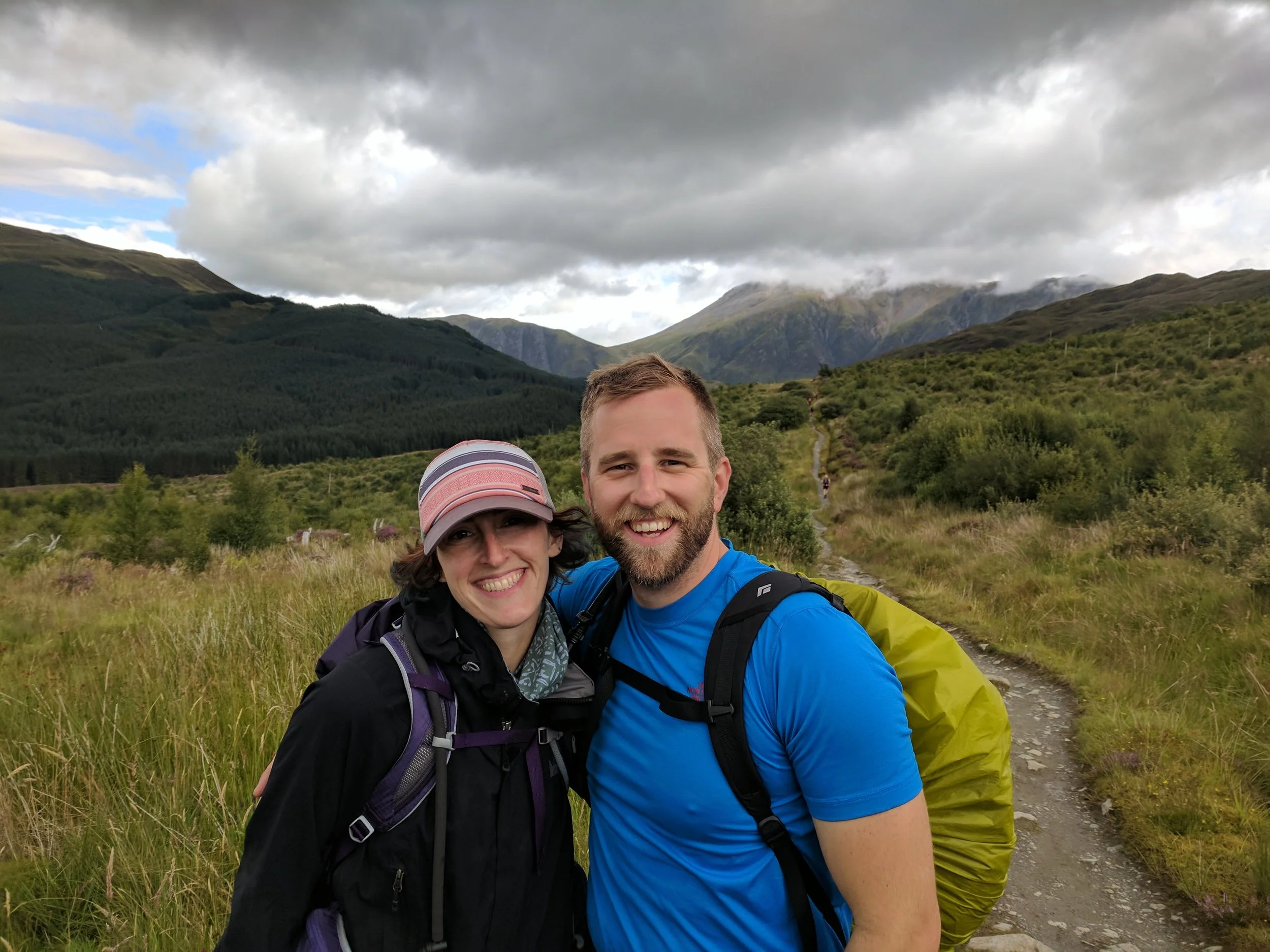 A smiling couple hiking on the West Highland Way trail, Scotland in a green mountainous landscape, with cloudy skies above.