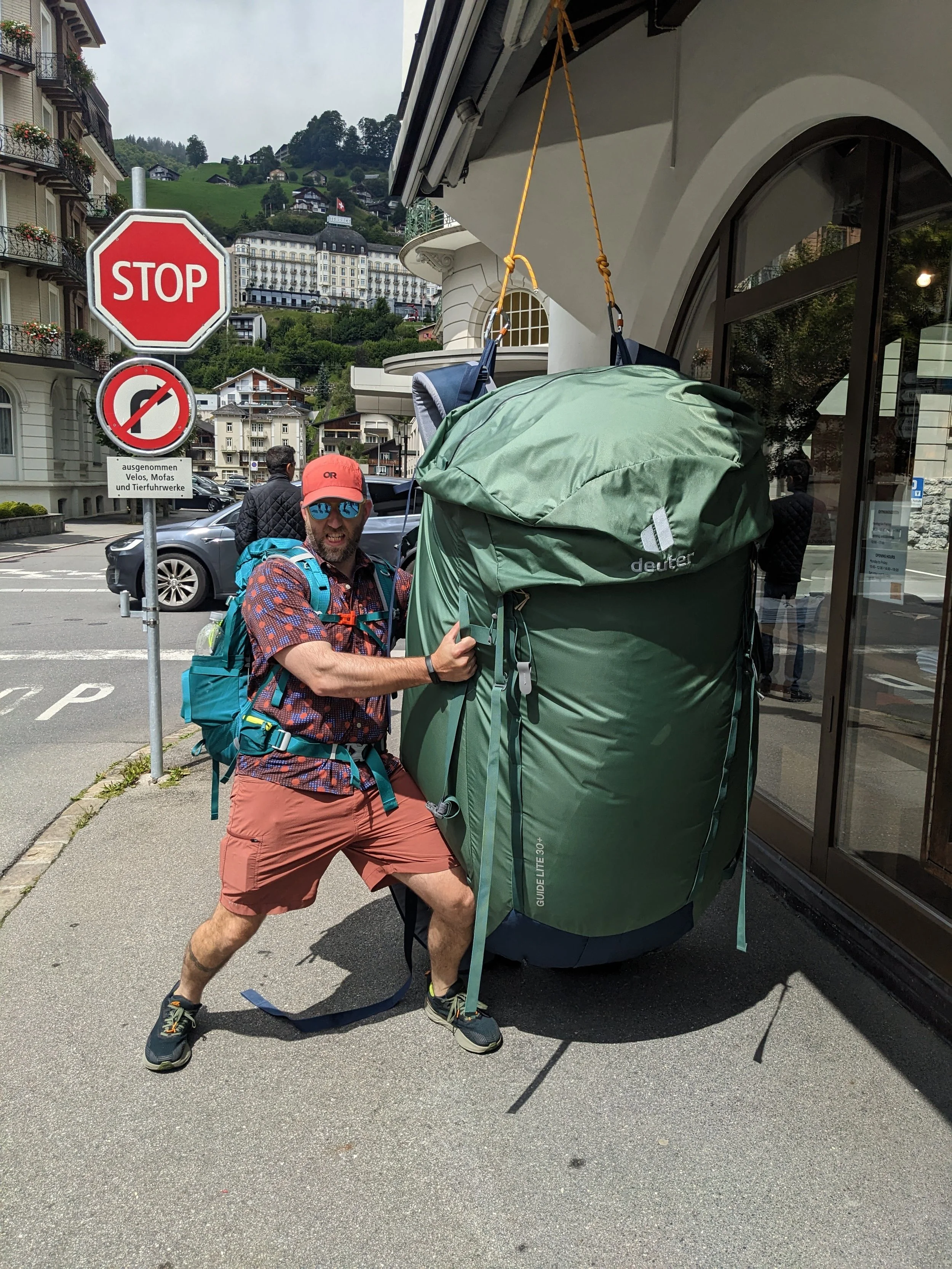 A man with a backpack and sunglasses posing next to a large green backpack outside a building on a city street in Switzerland on the Via Alpina 1 trail, with a stop sign and buildings in the background.