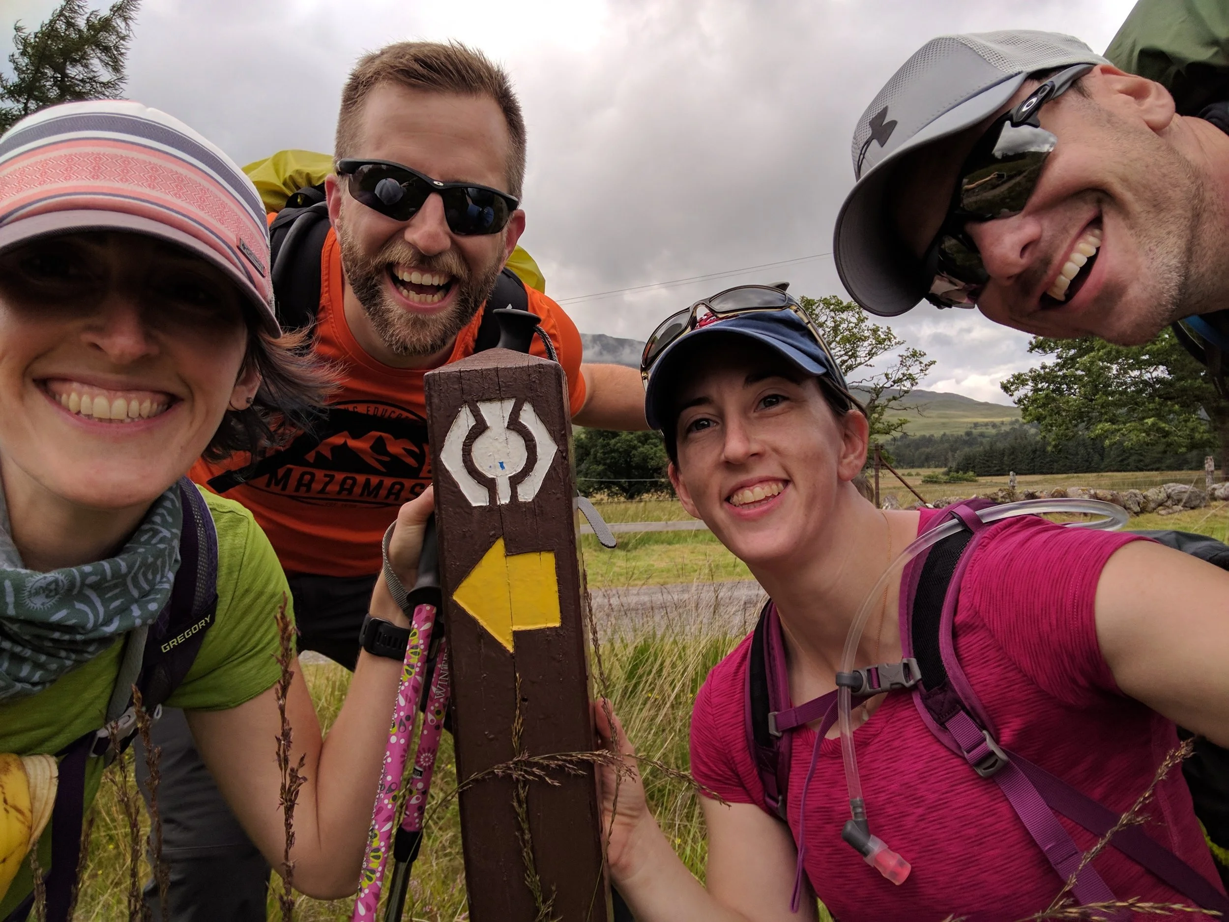 Five smiling hikers, three women and two men, pose together outdoors near a trail post with a yellow arrow and a circular emblem, in a grassy area with trees, hills, and cloudy sky in the background.