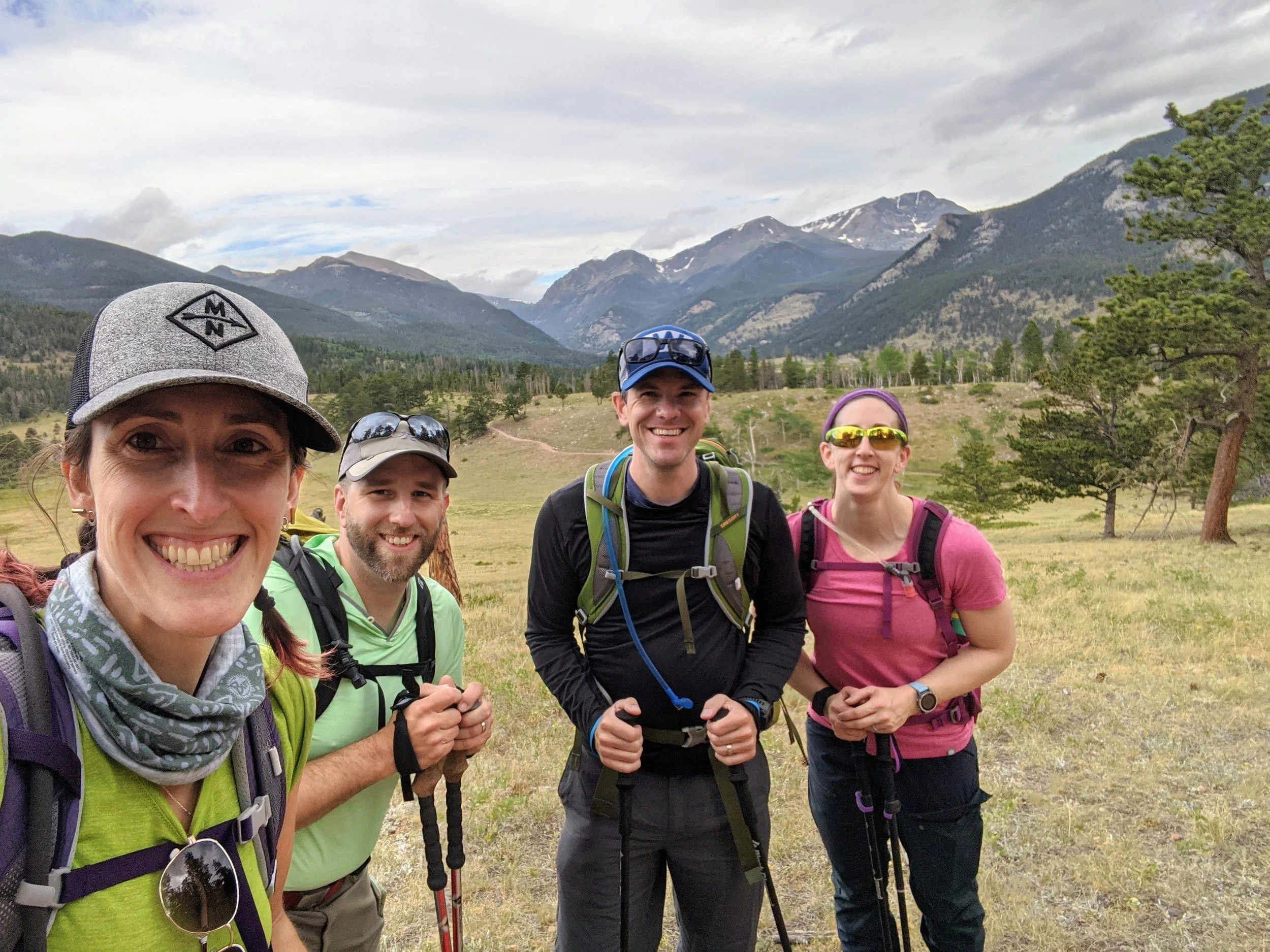 Group of four smiling hikers standing in a mountain meadow with mountains and trees in the background.