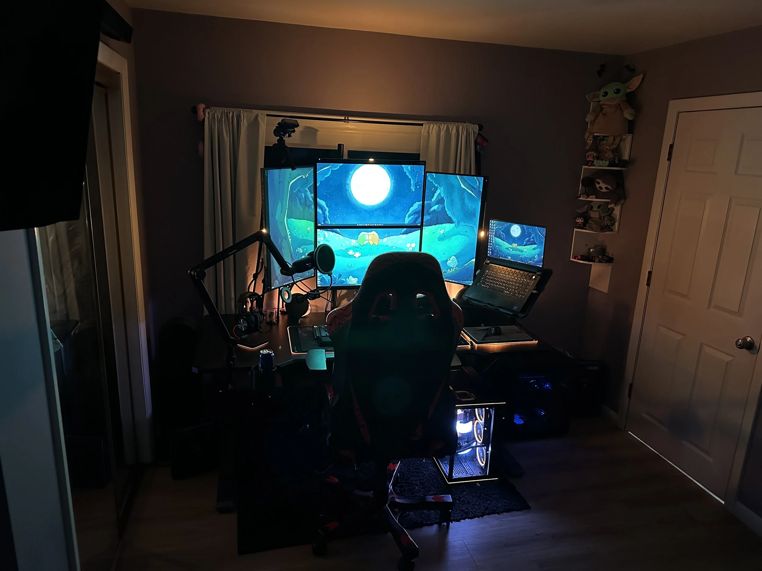 A computer desk setup with three monitors displaying a moonlit night scene, a microphone, headphones, a gaming chair, a computer case with visible cooling fans, and shelves holding plush toys.