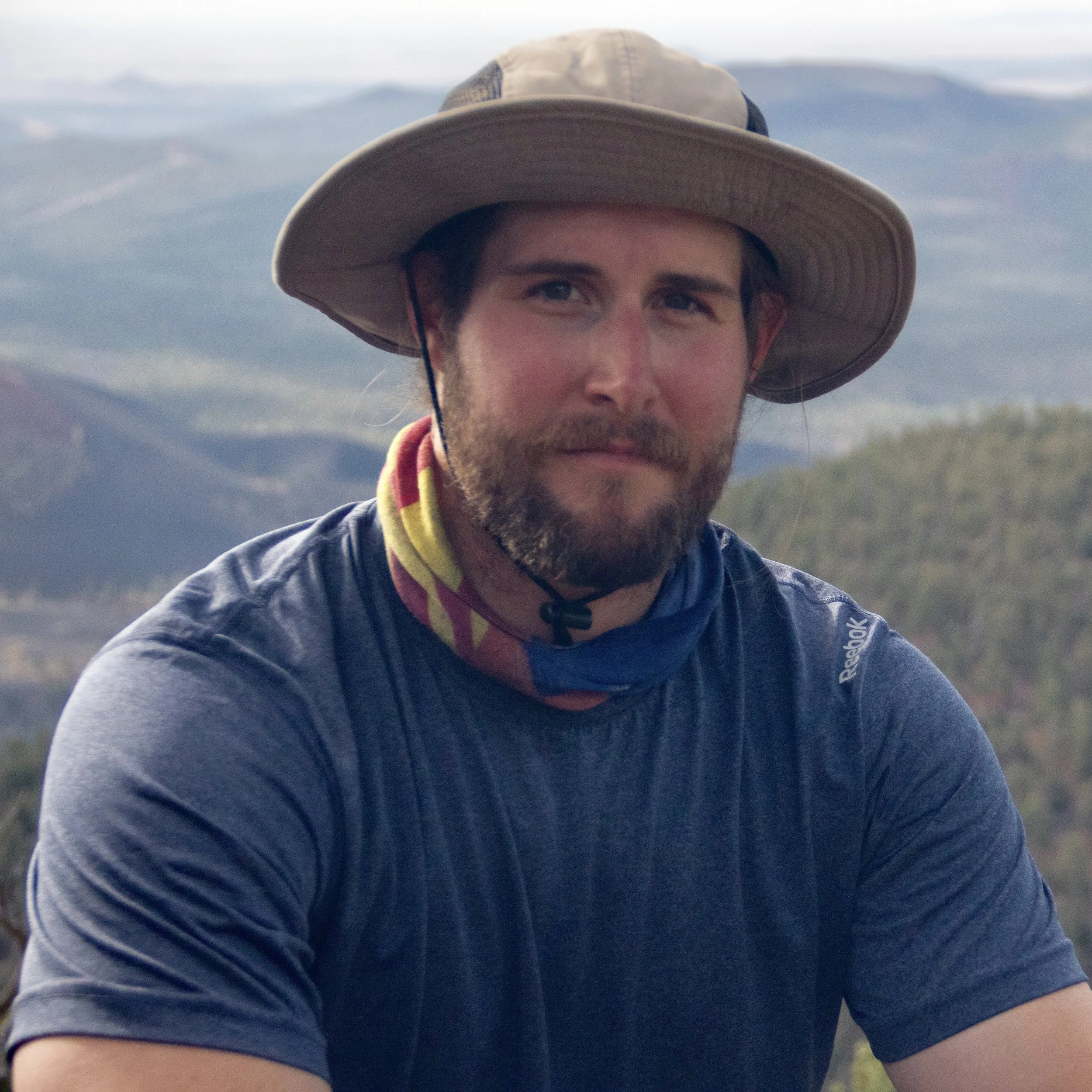 Man with a beard wearing a wide-brimmed hat, a blue T-shirt, and a colorful neck gaiter, sitting outdoors with mountainous landscape in the background.