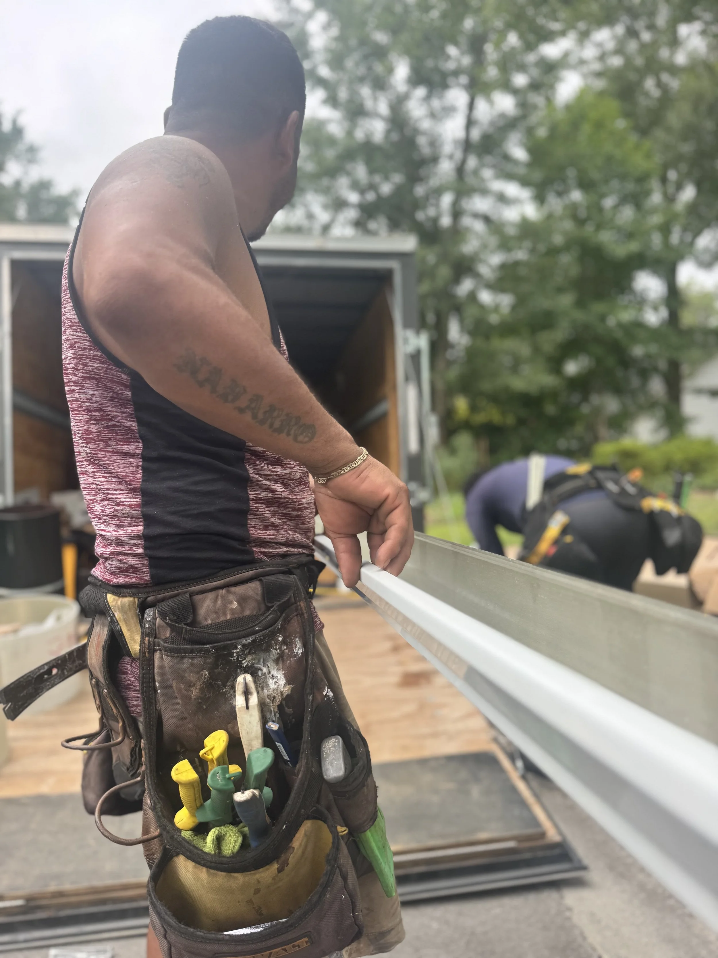 A construction worker in a tank top and tool belt measures a long piece of metal with a measuring tape indoors, with another worker in the background working.