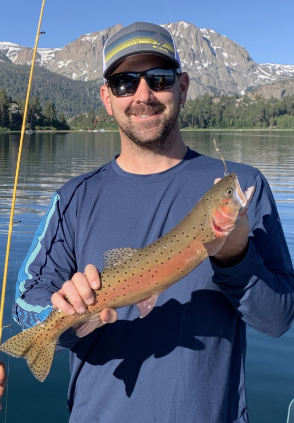 A man wearing sunglasses, a cap, and a long-sleeve shirt is smiling and holding a fish he caught. The background shows a lake with mountains and trees.