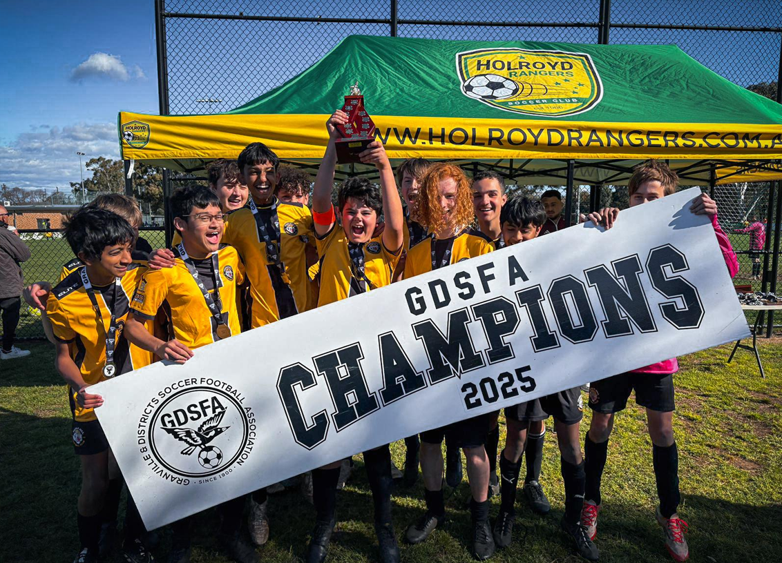Youth soccer team in yellow jerseys celebrating after winning a championship, holding a large banner that reads 'GDSFA Champions 2025.' They are outside on a soccer field with a green and yellow tent behind them.