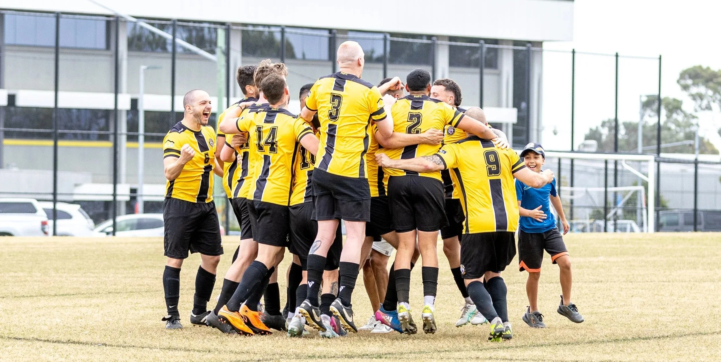 A team of soccer players in yellow and black uniforms celebrating together on the field, some hugging and smiling, with children and a soccer goal in the background.