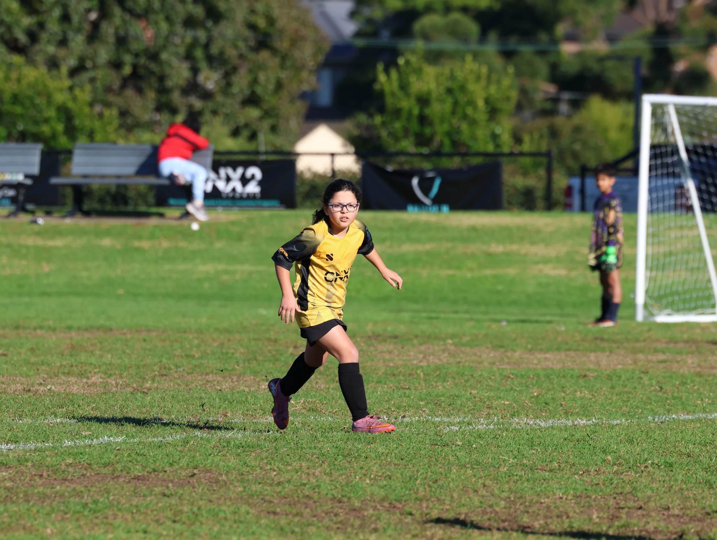 A girl playing soccer on a field with two other children in the background, a bench, trees, and a fence.