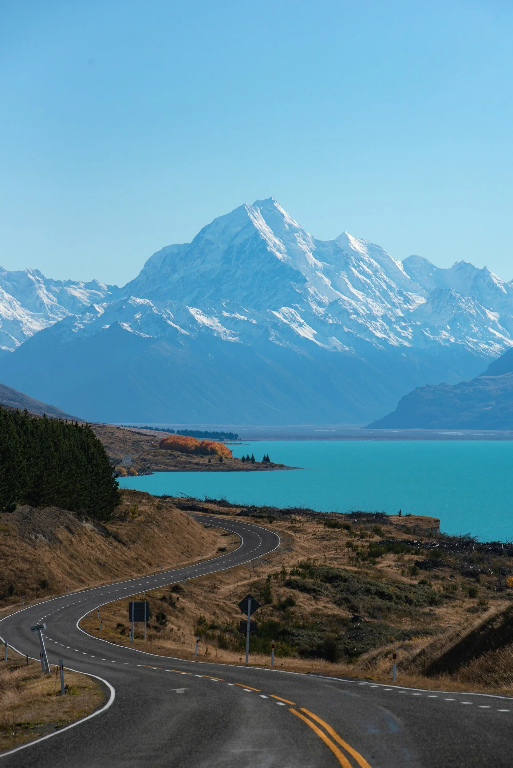 Winding mountain road with yellow dividing lines, leading toward snow-capped mountains and a clear blue sky.