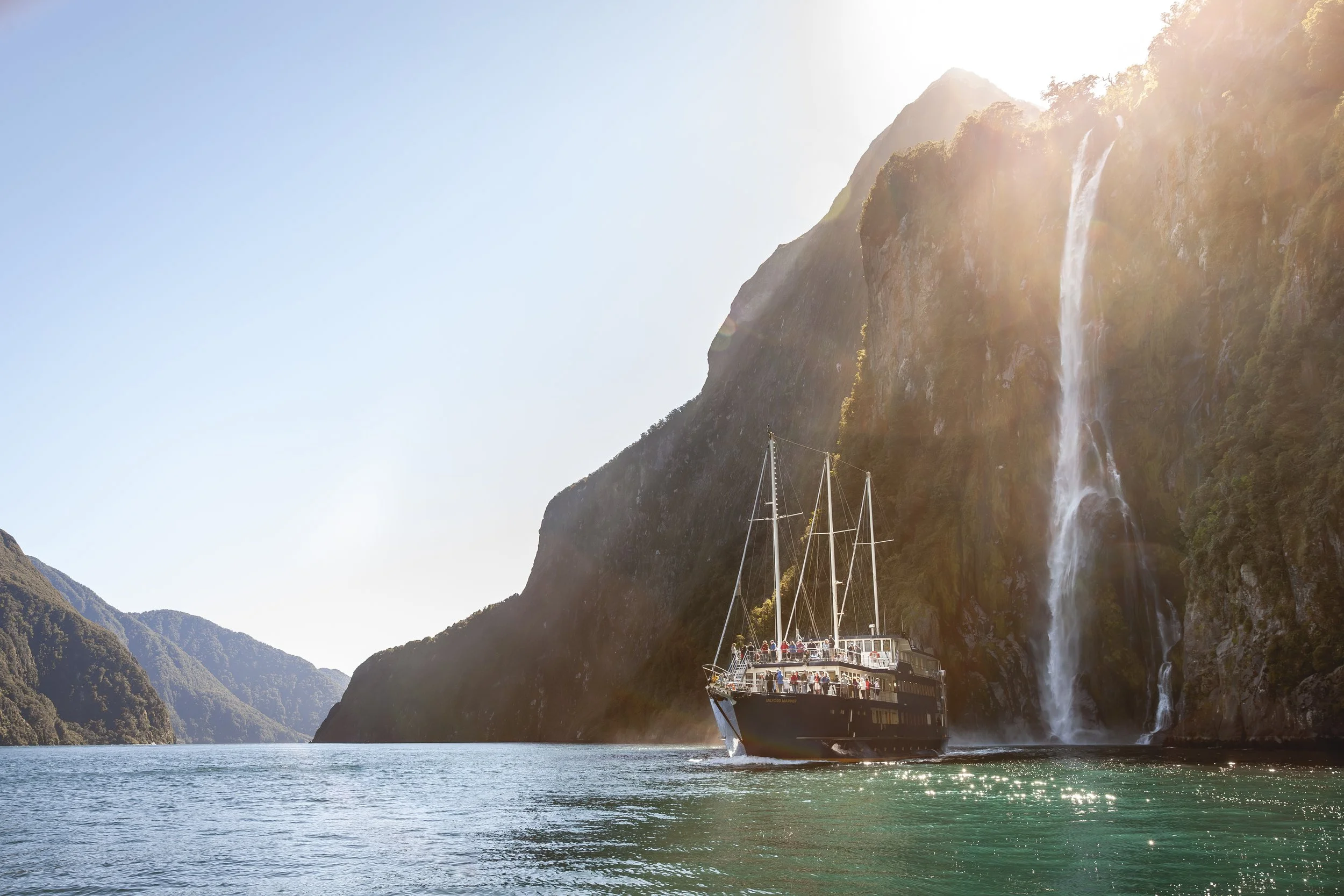 A boat sailing through a fjord with steep, green mountains on either side and a waterfall cascading down the mountain on the right, with sunlight shining from the top right corner.