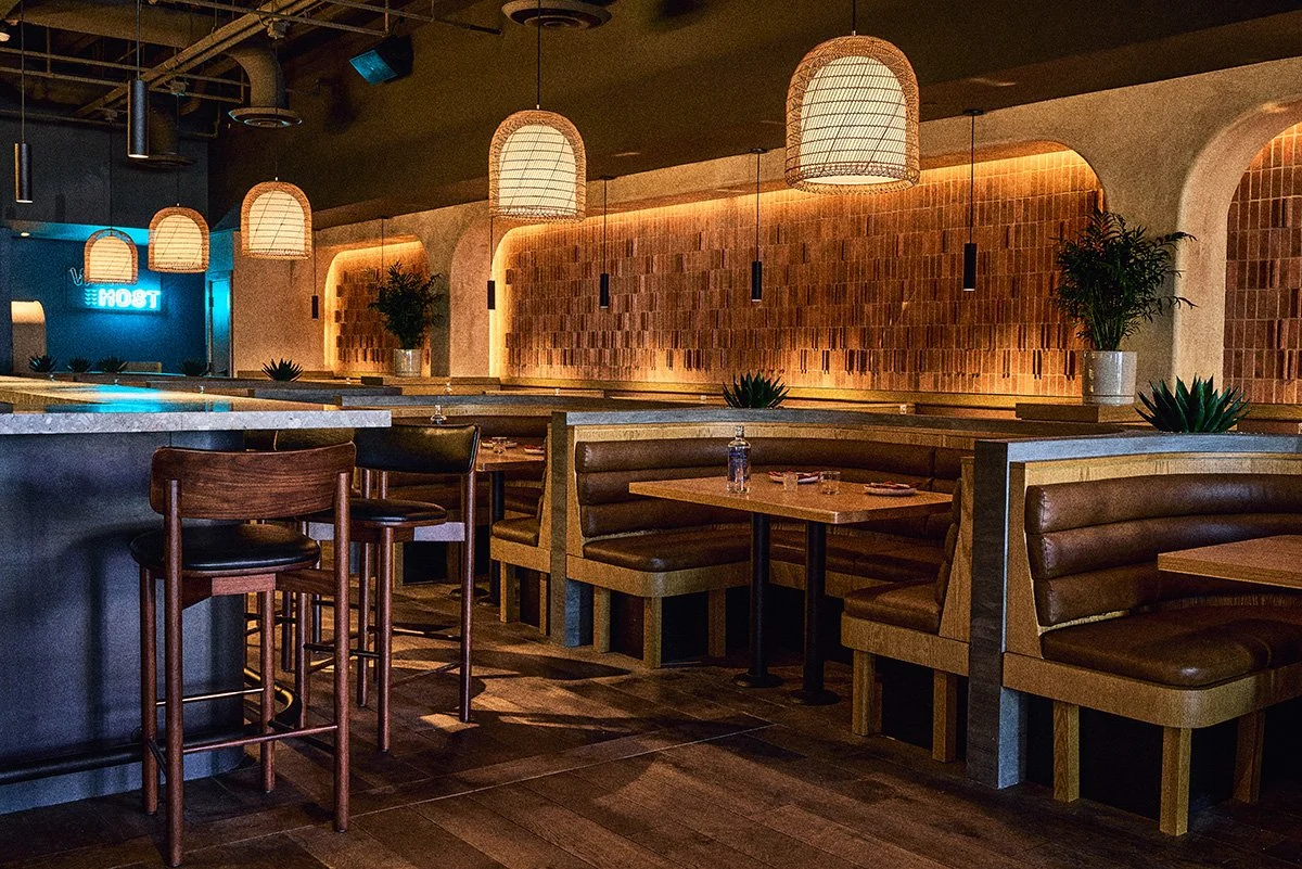 Interior of a modern restaurant with cushioned booth seats, wooden chairs, hanging wicker lamps, potted plants, and warm ambient lighting.