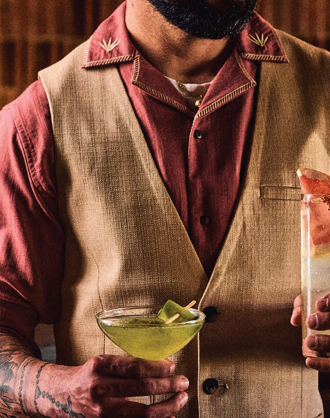 A man wearing a red shirt and beige vest holding a green cocktail glass with a lime garnish in one hand and a pink cocktail in a tall glass in the other.