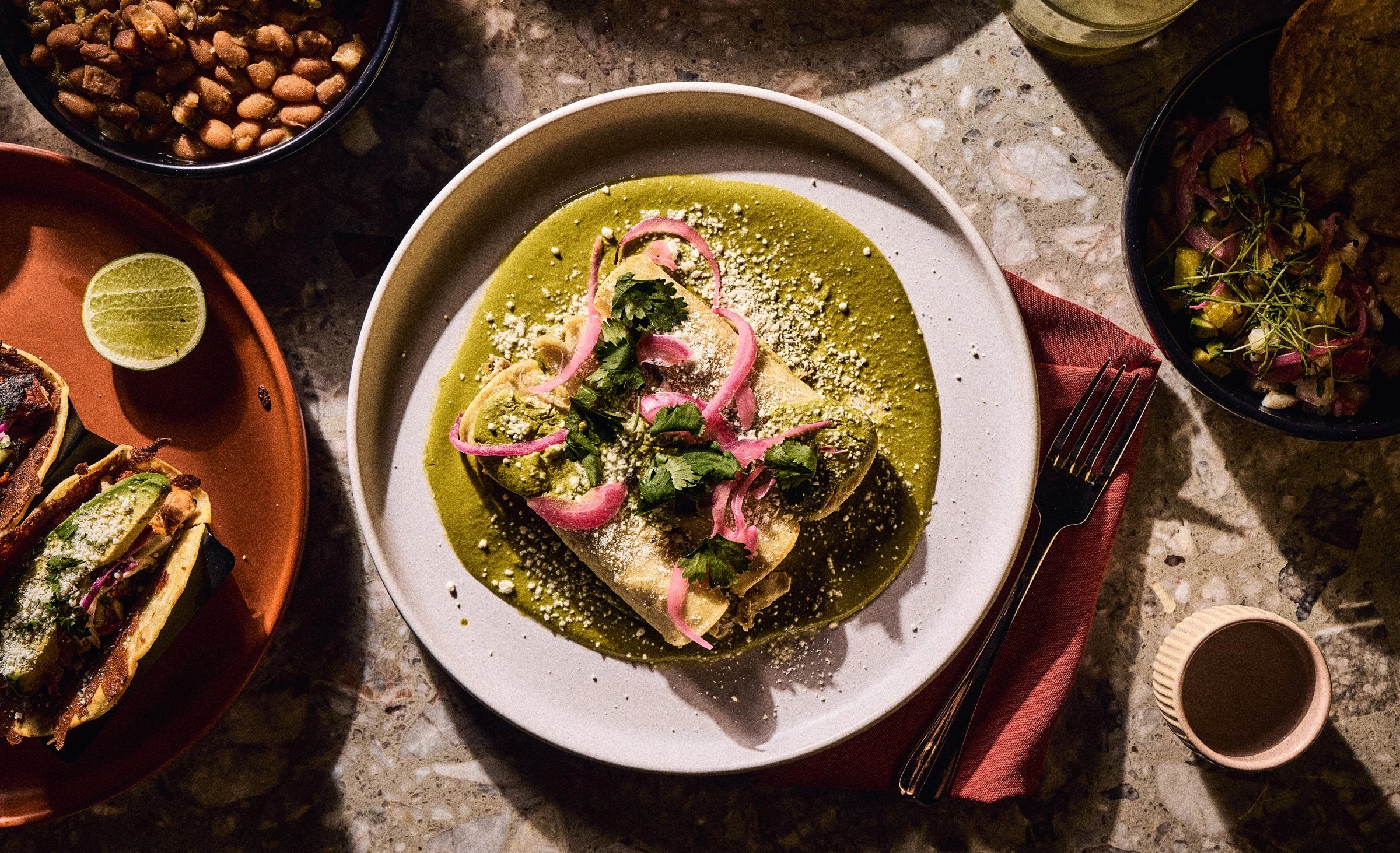 A white bowl of green sauce topped with cilantro leaves, sliced red onions, crumbled cheese, and a tortilla with a green filling. Surrounding dishes include a bowl of peanuts, a plate of sliced avocado, a quesadilla with guacamole on a red plate, and