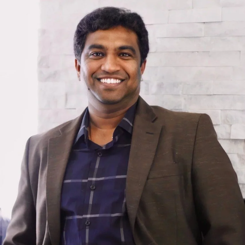A man in a brown blazer and a dark blue checkered shirt smiling, standing in front of a white brick wall.