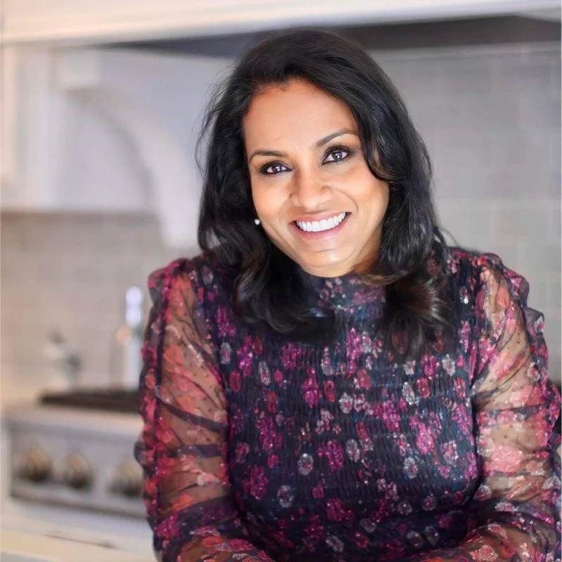 A woman with dark wavy hair, smiling, wearing a floral blouse with sheer sleeves, standing in a kitchen.