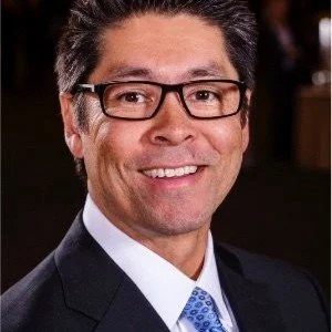 A smiling man wearing glasses, a dark suit, and a light blue tie.