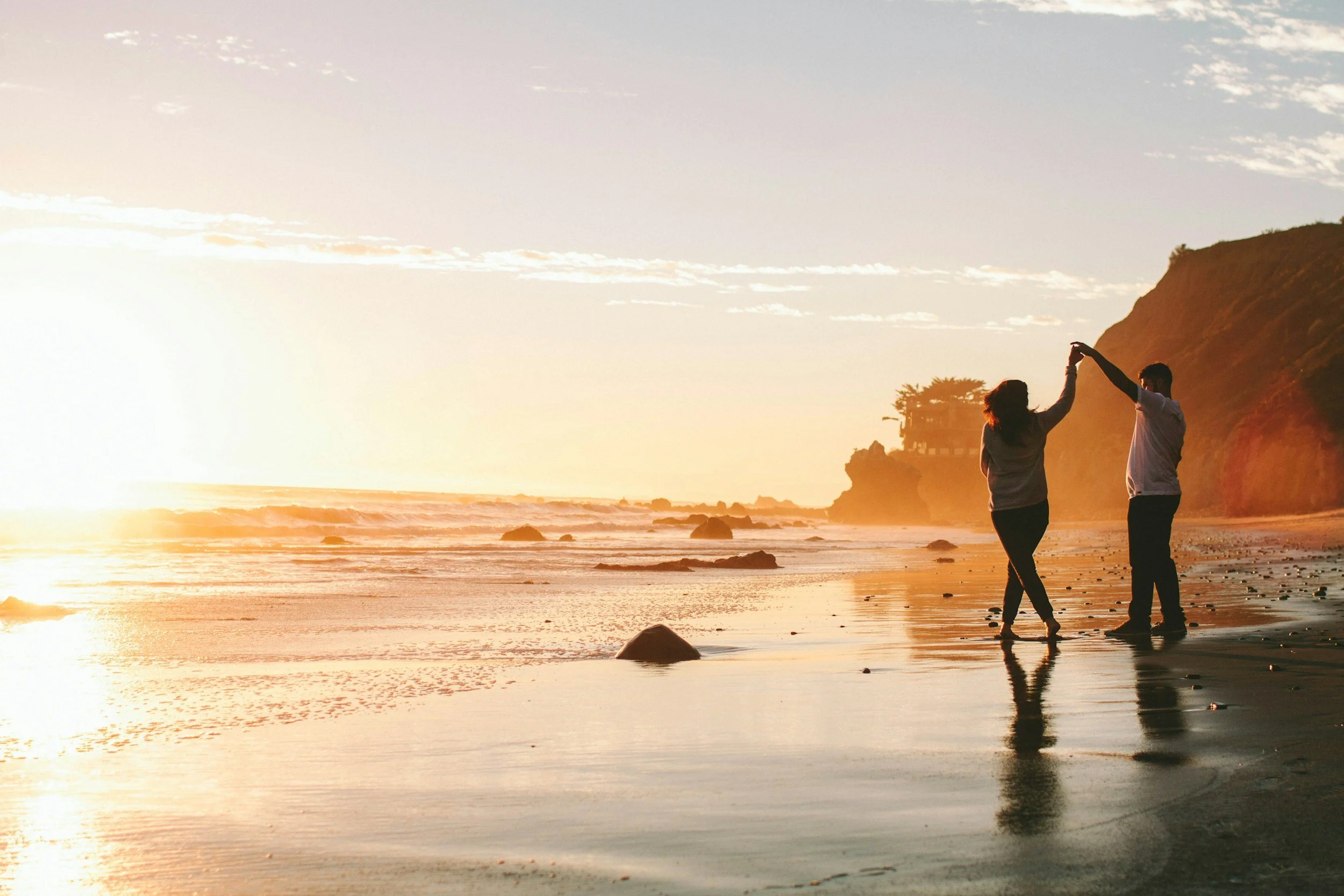 A couple holding hands and dancing on the beach at sunset, with a cliff and a small house in the background.